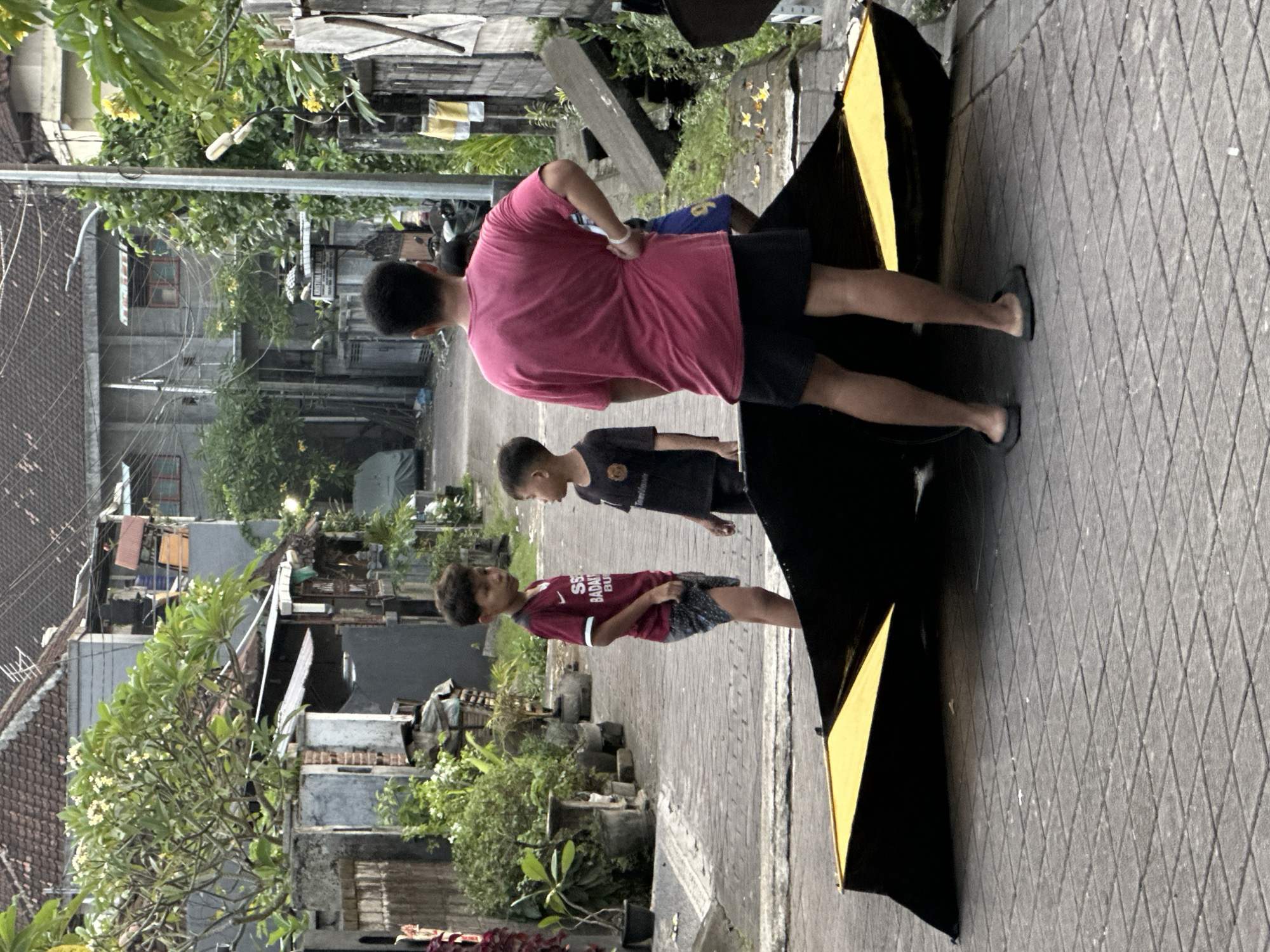 The image captures a street scene in North Kuta, Indonesia, featuring three males standing on a paved walkway. The adult male, closest to the viewer, wears a pink shirt and black shorts, his back to the camera. Two younger boys stand nearby, one in a dark T-shirt and the other in a burgundy shirt and patterned shorts. A black and yellow object stands beside them. The background includes buildings and vegetation lining the walkway. The perspective is from an elevated position, looking down on the scene.