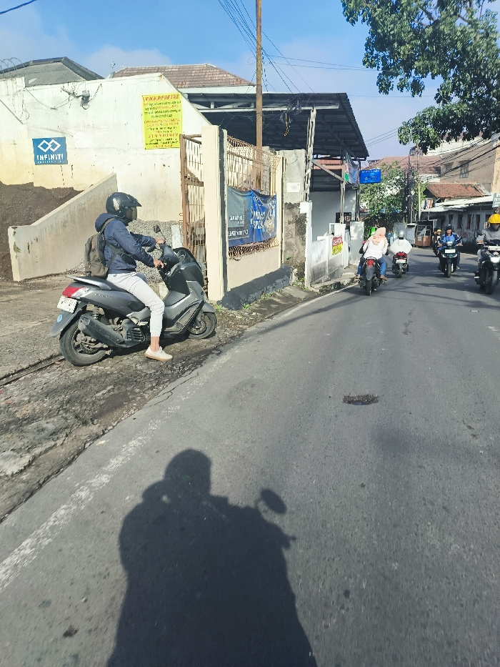 The image shows a street scene in Bandung, Indonesia, taken at coordinates -6.892, 107.629. A man in a helmet and a dark jacket is riding a motorcycle on a paved road with a white stripe on the right side. He is wearing white pants and white shoes. The motorcycle is a silver scooter with a black seat. Behind him are several other motorcycles and people. Some are also riding motorcycles, while others are walking. The street is flanked on the left by a building with a gray wall, and on the right by a row of buildings with a variety of colors and textures. The image is taken from a low angle and the perspective is slightly elevated. There are several shadows on the ground. The sky is a clear blue with a few white clouds.