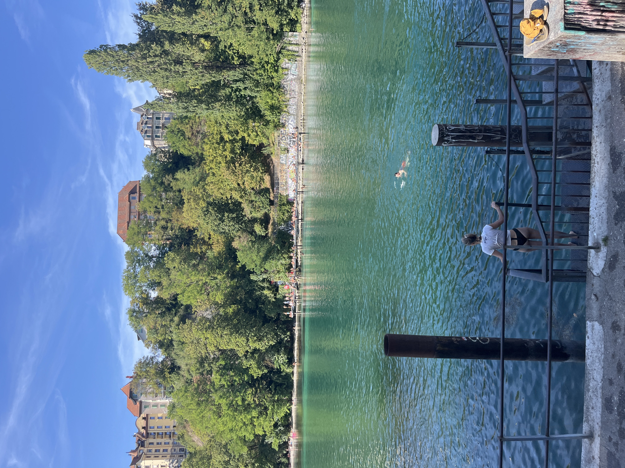 The image depicts a sunny day at the Jetée des Pâquis in Geneva, Switzerland, a popular waterfront area. In the foreground, a woman wearing a white t-shirt and black bikini bottoms stands at the top of a metal staircase leading down to the turquoise water. She is looking towards the water, holding onto the railing. Further into the water, two individuals are swimming. The middle ground shows a wooden pier extending into the lake, with a crowd of people sunbathing and enjoying the water. Along the embankment behind the pier, trees and buildings are visible, with a clear blue sky overhead. The water is calm and reflects the sunlight.