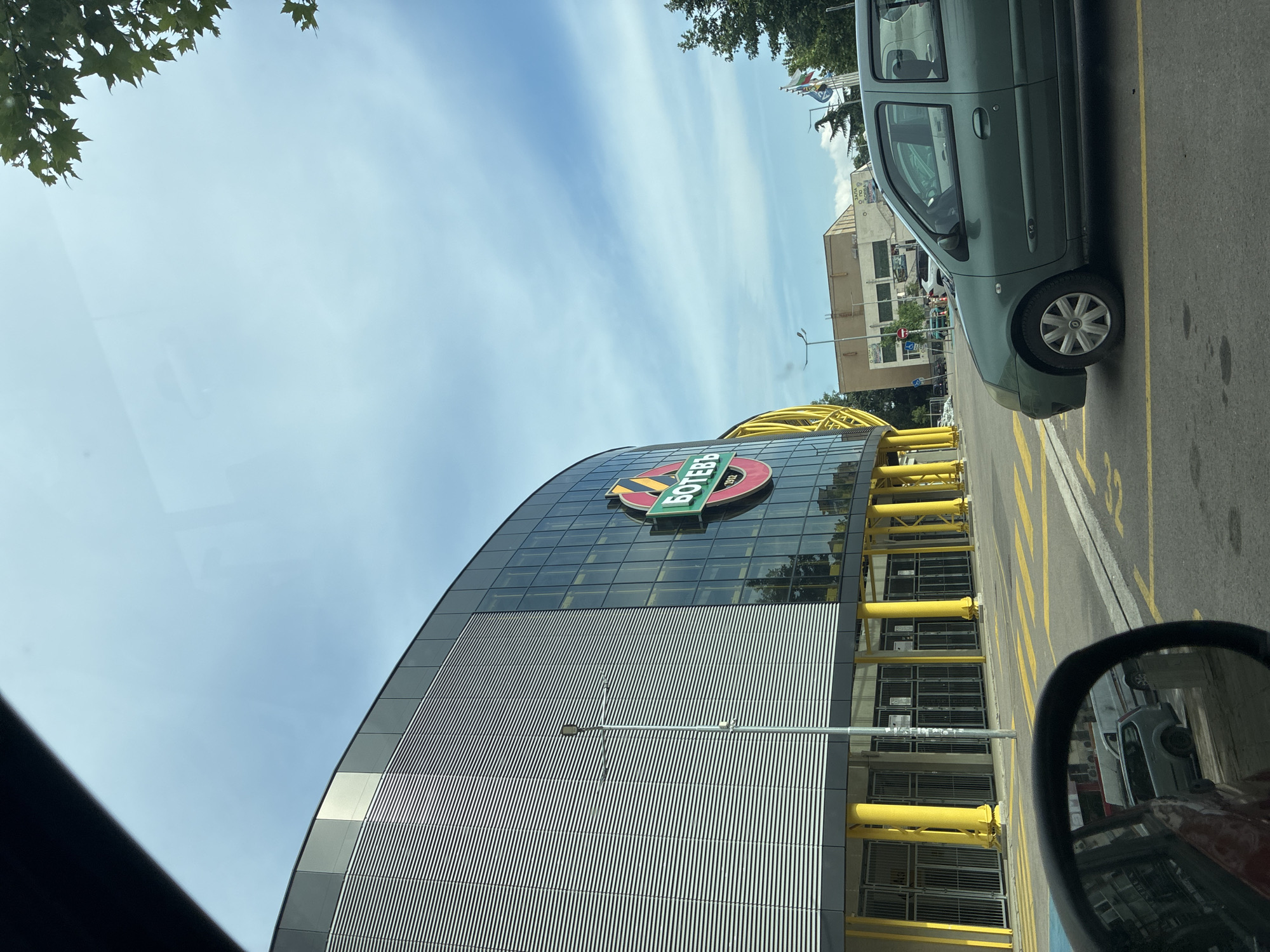 The image, taken from inside a vehicle, captures a modern building under a bright blue sky with wispy clouds. The perspective suggests the original image was rotated 90 degrees clockwise.
The central subject is a large, contemporary building featuring a curved facade, with its upper section clad in dark glass panels and the lower part in horizontal metallic ridged panels. A prominent circular logo is affixed to the glass facade. This logo consists of a red circle with a green horizontal banner across it, displaying the Cyrillic text "БОТЕВЪ" (Botev) in green letters. Below the banner, within the red circle, the year "1912" is visible. Along the base of the building, a series of bright yellow cylindrical pillars or support structures create a striking architectural element. A lamppost stands in front of the building.
On the right side of the frame, a dark green compact car, possibly a minivan or small SUV, is parked on asphalt. Yellow lines indicating parking spaces are visible on the asphalt, with one marking clearly showing the number "12". The bottom right corner shows a portion of a car's side mirror, which reflects another vehicle (light-colored) and parts of the building.
Green tree foliage is visible in the top left corner and further in the background on the right. In the distance, behind the parked car, other urban buildings and several flags on poles can be faintly seen. The scene is illuminated by daylight, indicating clear, sunny weather. There are no people visible, and no specific activity is taking place. The visible text "БОТЕВЪ 1912" strongly suggests the building is associated with PFC Botev Plovdiv, a football club, consistent with the location context of Plovdiv, Bulgaria.