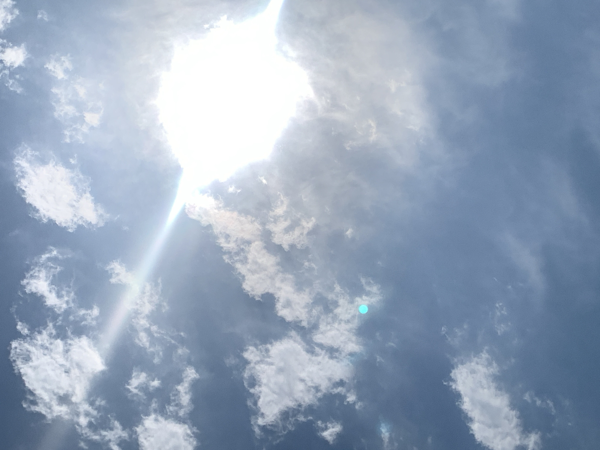 The image depicts a bright, cloudy sky with the sun prominently featured. The sun's rays create a lens flare effect, diagonally bisecting the frame from the upper left to the lower right. The sky is a deep blue, punctuated by scattered white clouds of varying sizes and densities. One small, teal-colored circular anomaly is visible in the middle left portion of the image. The scene appears to be an outdoor, daytime view of the sky, characteristic of the weather in Khazar Raion, Azerbaijan. There are no people, subjects, objects, or discernible actions within the frame.
