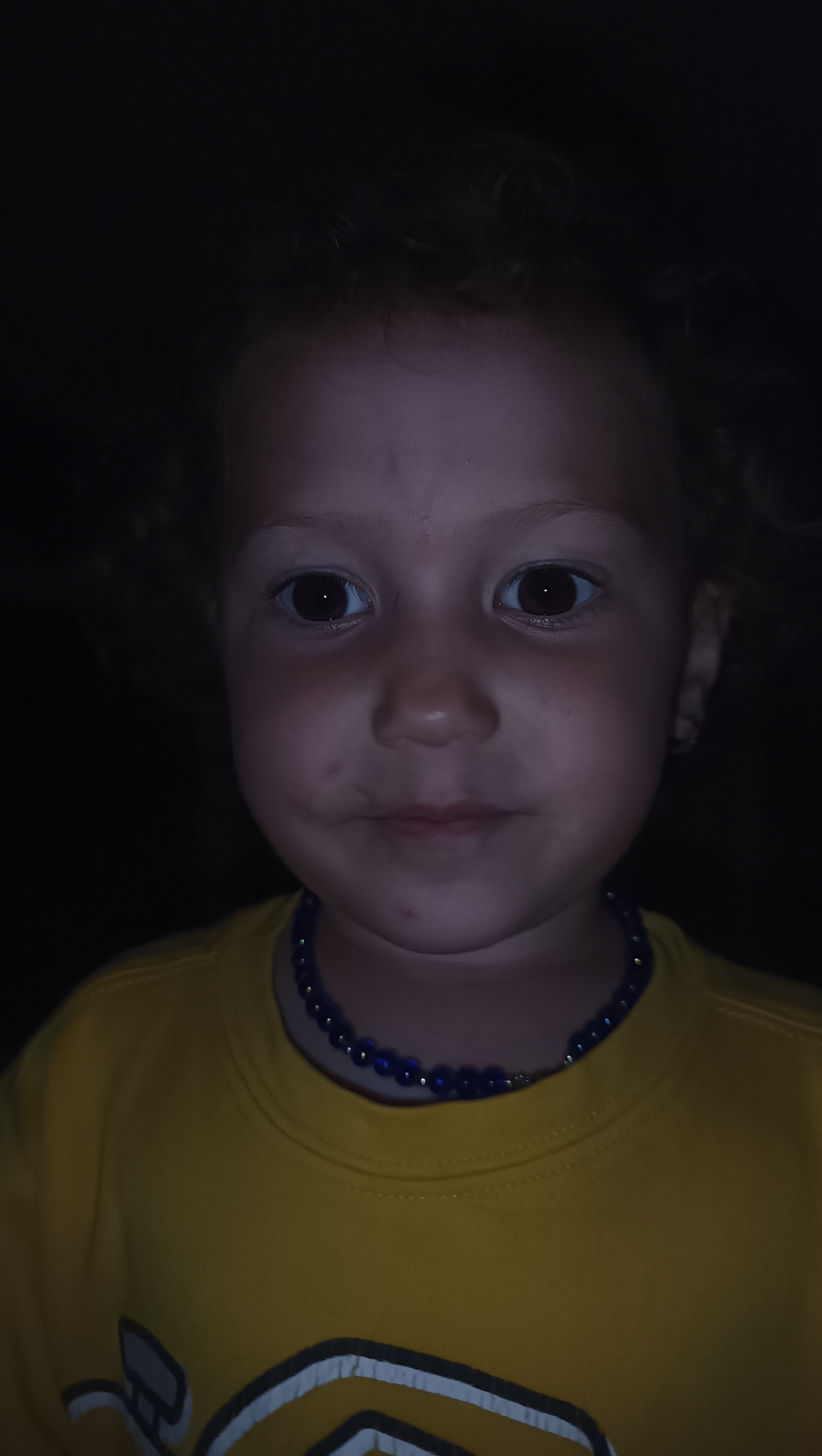 A close-up, head-and-shoulders shot of a young child, likely a girl, is presented in low light conditions. The child has curly brown hair, large brown eyes, and a fair complexion. She is wearing a yellow long-sleeved shirt and a dark blue beaded necklace. The background is dark and indistinct. No specific location details can be determined from the image, and therefore no location in the city of Karsha, Russia can be provided.