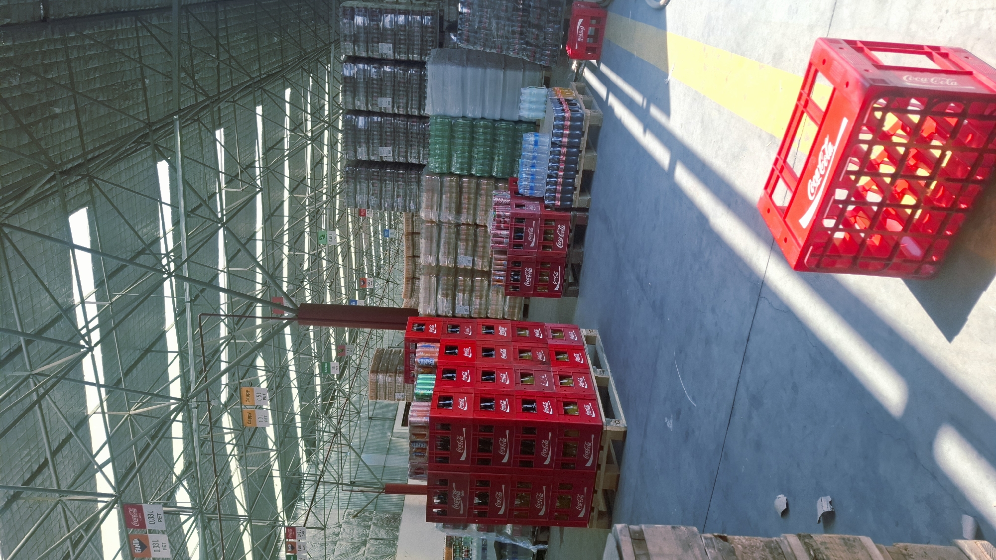 The image depicts the interior of a large warehouse, likely a distribution center, filled with stacks of beverages. In the foreground, several red Coca-Cola crates are visible, some filled with bottles and others empty. Pallets laden with cases of bottled and canned drinks, primarily Coca-Cola products, are stacked throughout the warehouse. Various types of bottles, including clear and green glass, are also present. The ceiling is a complex metal structure with translucent panels allowing natural light to filter in, creating distinct shadows on the concrete floor. Hanging signs indicating product details like "0.33 L PET" and brand names such as "Coca-Cola" and "Fanta" are suspended from the ceiling. The overall impression is one of organized storage and inventory of beverages. The location is specified as Fergana City, Uzbekistan.