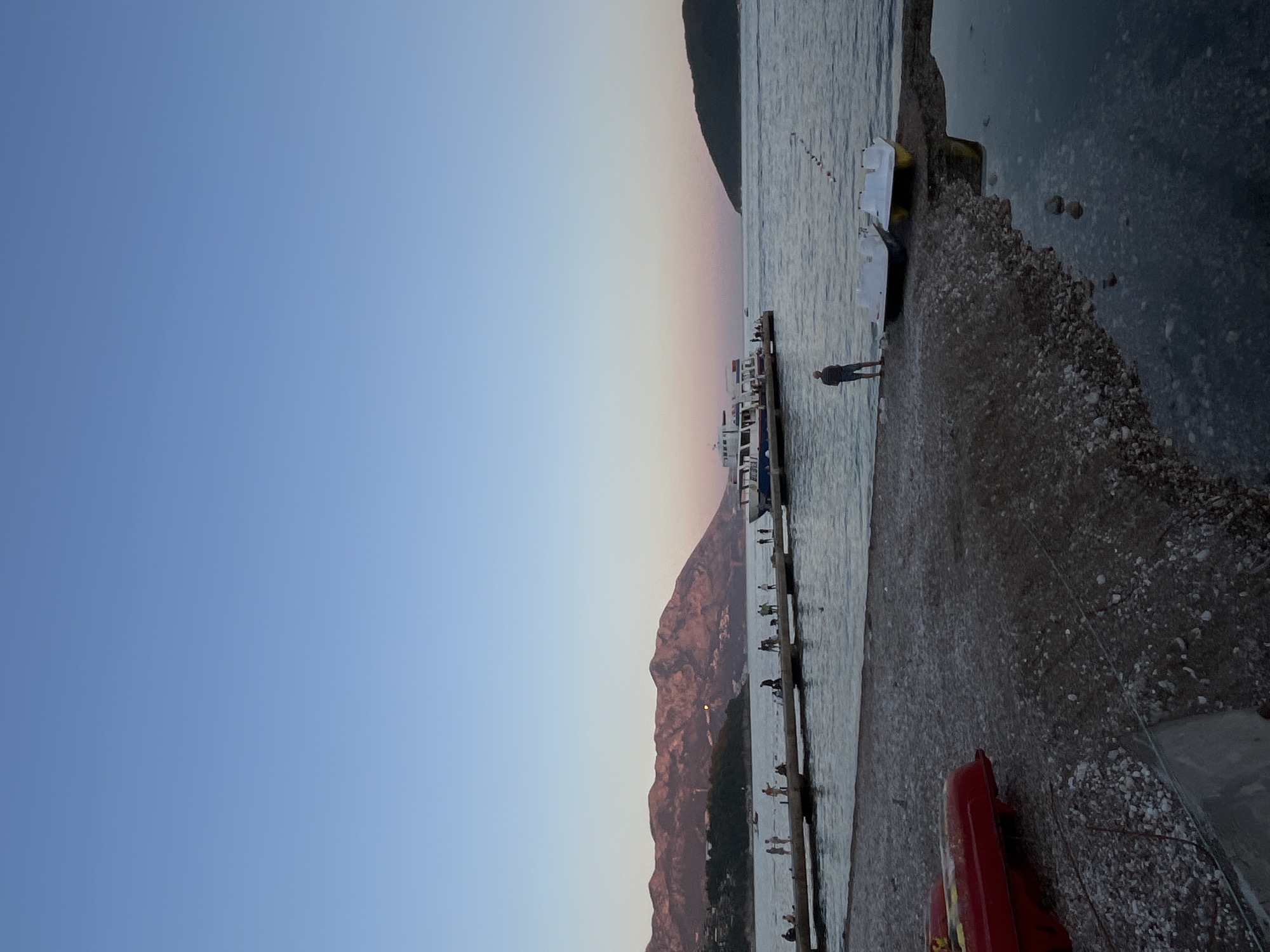 This image depicts a sunset scene at a coastal area in Budva, Montenegro. In the foreground, a pebbly beach is visible, with two red pedal boats partially submerged in the shallow water at the left edge. Further back, a person stands on the shore, facing two white pedal boats docked at the water's edge. A long, concrete pier extends into the calm sea, where several people are seen walking and standing. Two passenger boats are docked at the pier. In the background, a range of mountains with pinkish hues from the setting sun stretches across the horizon, and a smaller, dark island is visible to the right. The sky transitions from a clear blue at the top to a soft pink and orange gradient near the horizon.