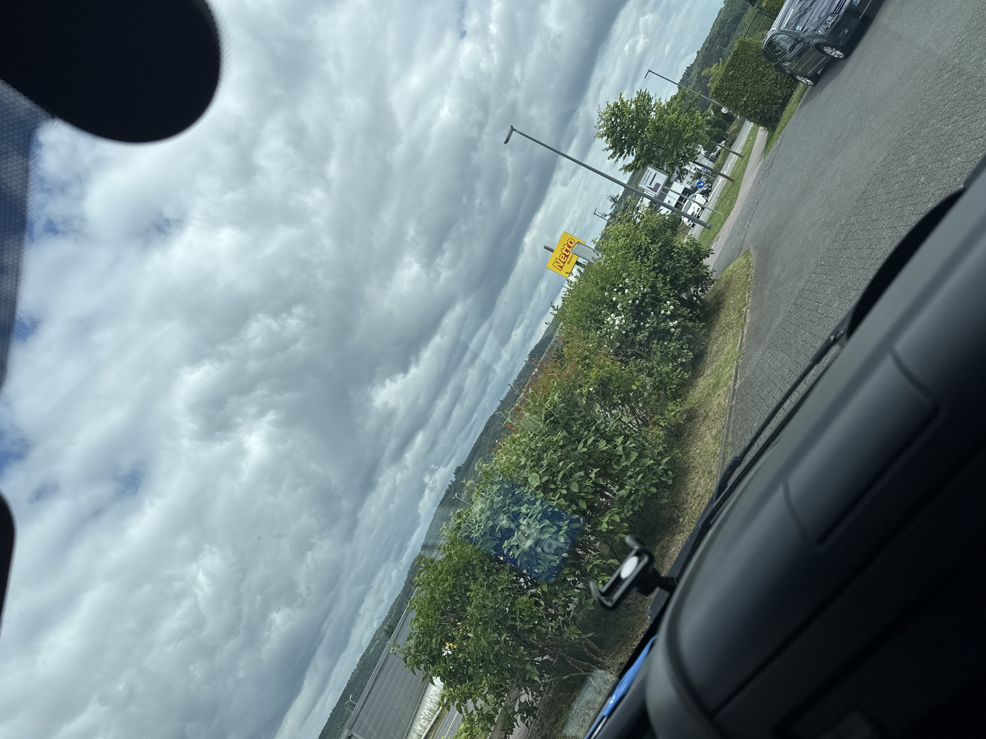 The image, taken from inside a vehicle looking out, depicts an overcast sky with dramatic clouds. In the middle ground, a lush green area with trees and bushes borders a paved parking lot. A bright yellow sign with the word "Netto" is visible on the left side of the road, indicating a supermarket. Several cars are parked in the lot. A dark-colored car is parked on the right. The scene suggests a commercial area, possibly in the city of Ulmen, Germany, given the presence of a Netto supermarket, which is a common chain in Germany. The environment appears to be a suburban or semi-rural setting with a mix of natural elements and man-made infrastructure.