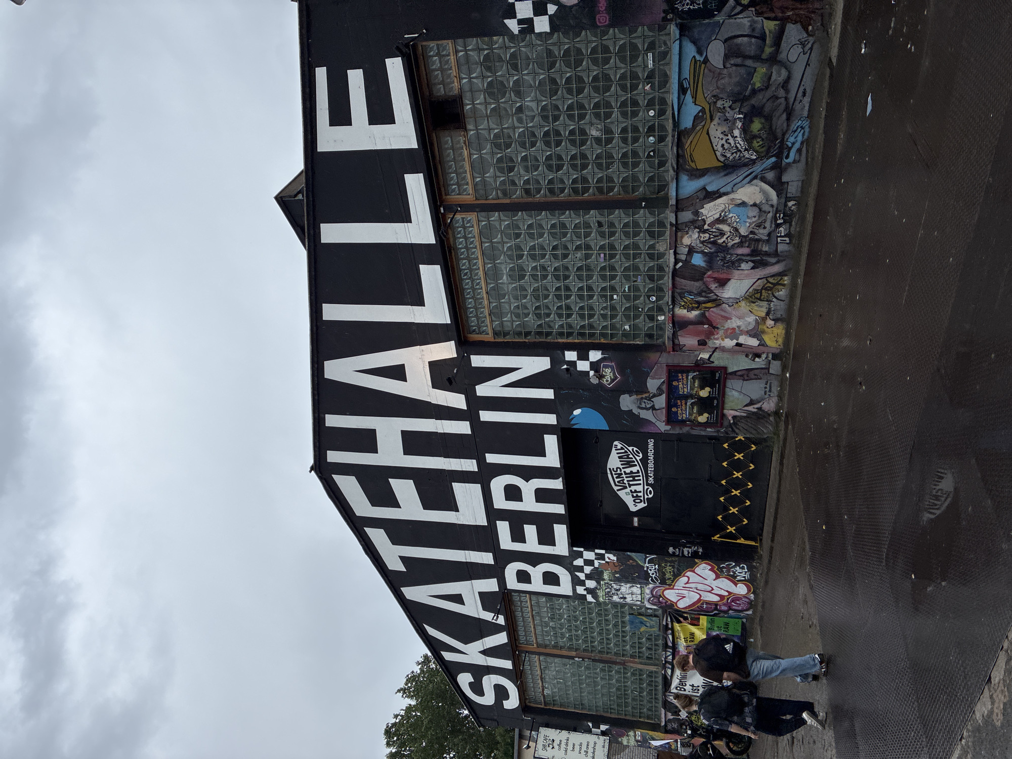 The image shows the "Skatehalle Berlin," a well-known indoor skatepark located at Revaler Straße 9, Berlin, Germany. The building features large white lettering across its black facade that reads "SKATEHALLE BERLIN." The entrance is a black gate with a "Vans Off The Wall Skateboarding" logo. The exterior walls are heavily decorated with graffiti and murals. In the foreground, two individuals are walking away from the camera, one wearing a black backpack and the other a brown backpack. The ground is wet and appears to be a metal grate or textured surface, reflecting the overcast sky. The overall environment suggests an urban setting with a focus on skateboarding culture.