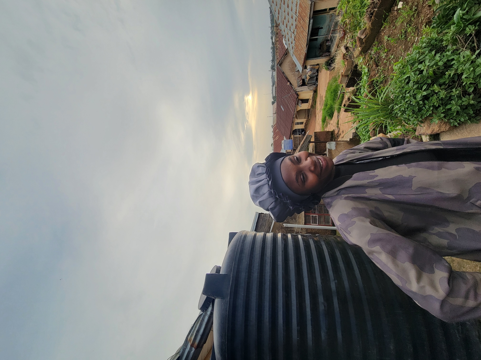 The image depicts a woman in a camouflage jacket and a grey head covering, smiling and looking to her right. She is positioned in the foreground, with a large black water tank to her left. Behind her, a view of a residential area in Jos, Nigeria, is visible. The houses have corrugated iron roofs, and the sky is overcast with a hint of sunlight breaking through. The environment appears somewhat rural, with some greenery and dirt paths between the buildings. The woman's clothing and the surroundings suggest a casual outdoor setting.