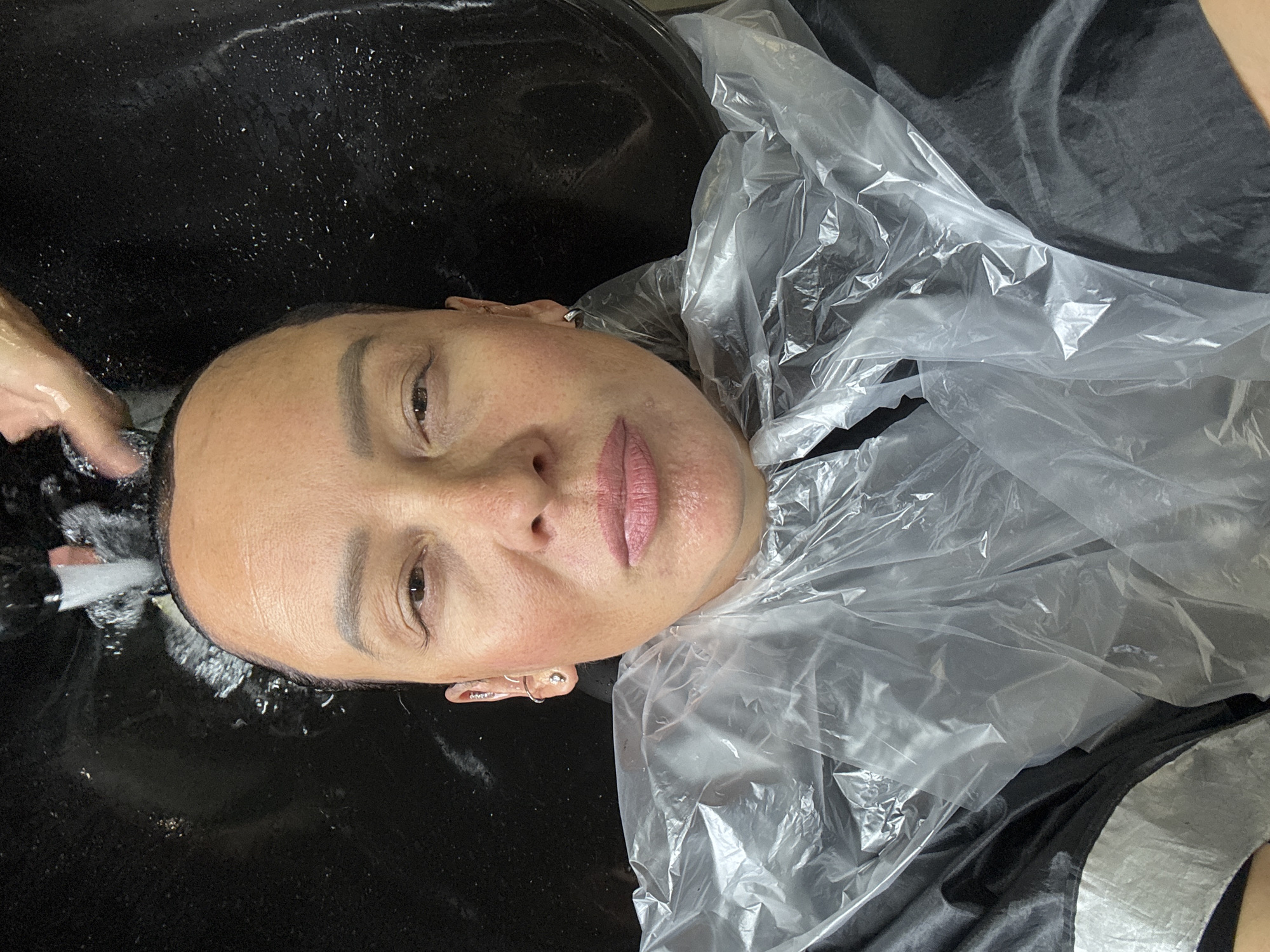 A woman is at a hair salon in Chaikovsky, Russia. She is lying on her back in a black hair-washing sink, having her hair washed. She is wearing a clear plastic cape and a black gown. A hand is visible, presumably that of the salon employee, washing the woman's hair.