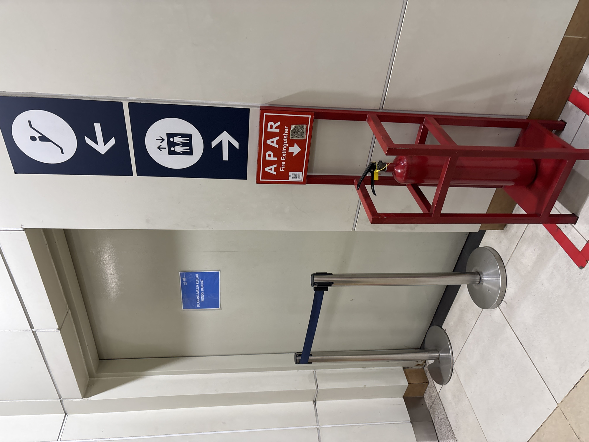 The image shows a hallway in what appears to be a public building, possibly an airport or train station, given the signage and crowd control barriers. The setting is indoors, with tiled flooring and light-colored walls.
The primary subjects are directional signs and a fire extinguisher. On the left, there are two dark blue signs with white pictograms and arrows. The first sign shows a person in a diving pose with a downward arrow, indicating a downward direction. The second sign depicts multiple figures with upward and downward arrows, suggesting movement of people in both directions. To the right of these signs, a red metal stand holds a red fire extinguisher labeled "APAR Fire Extinguisher" in white text, with a downward arrow and a QR code.
In the foreground, two silver stanchions with blue retractable belts are set up, suggesting a queue or restricted access area. A door is visible on the left behind the stanchions, with a blue sign attached to it. This sign contains text that appears to be in Indonesian, with a warning that access is prohibited except in emergencies.
The overall scene is one of an organized public space with clear indications for movement and safety equipment. The lighting suggests it is daytime, but no external weather conditions are visible. The location cues, such as the "APAR" label (common in Indonesia for fire extinguishers) and the Indonesian text on the door sign, point to this being in Central Jakarta, Indonesia. There are no people actively engaged in any specific activity, but the presence of directional signs and barriers implies ongoing foot traffic or controlled entry.