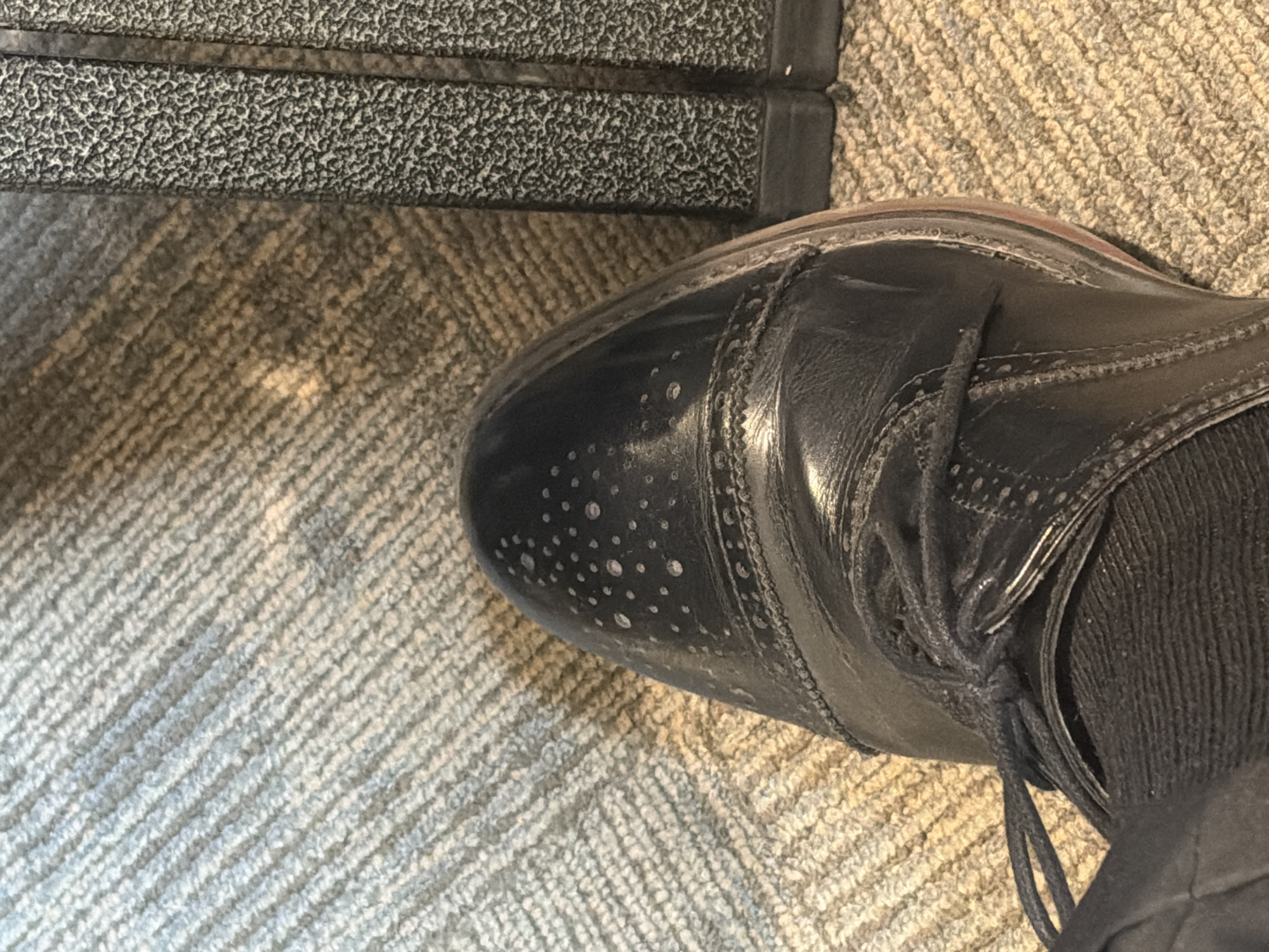 An individual's right foot is positioned on a carpeted floor. The foot is clad in a black leather dress shoe with visible brogue detailing and perforations on the toe cap. The shoe is laced with black laces. A black sock is visible above the shoe. The carpet appears to be a textured loop pile in shades of beige and gray. To the right of the foot is a dark gray, textured object, possibly furniture or a piece of equipment. The scene suggests an indoor setting. There is no information available to determine the location within the city of Plymouth, United States of America.