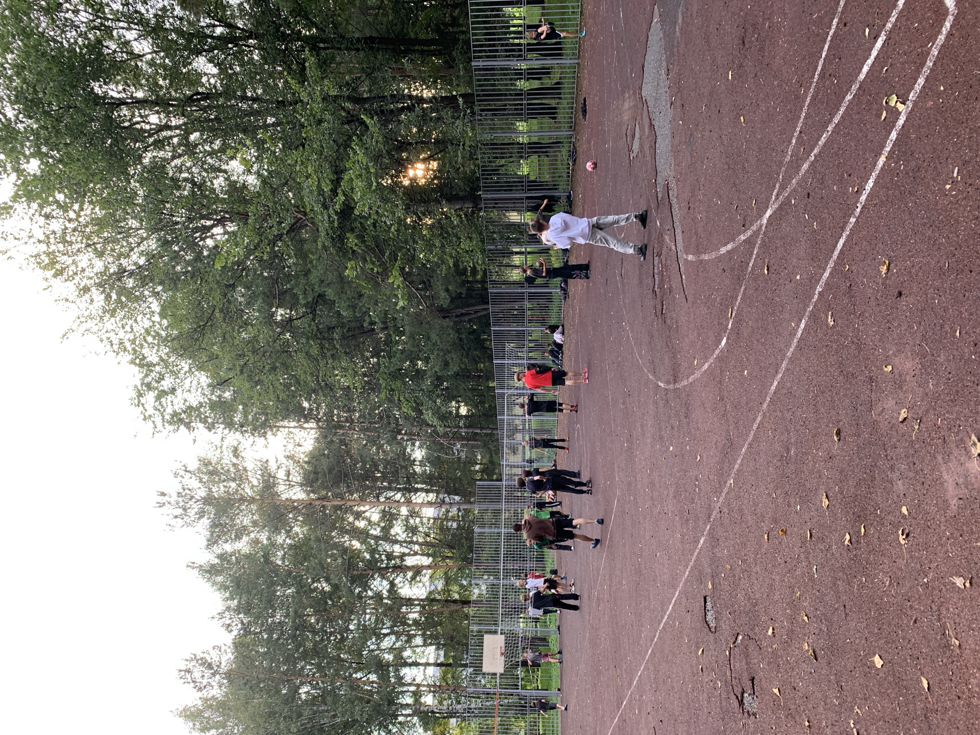 A group of young people are playing on a red, outdoor basketball court surrounded by a metal fence and tall trees. The court has white markings, including a three-point line and a center circle. There are several patches of damage and debris on the court surface. In the background, the sun is setting through the trees, casting a warm glow. The location is Prokopyevsk, Russia.