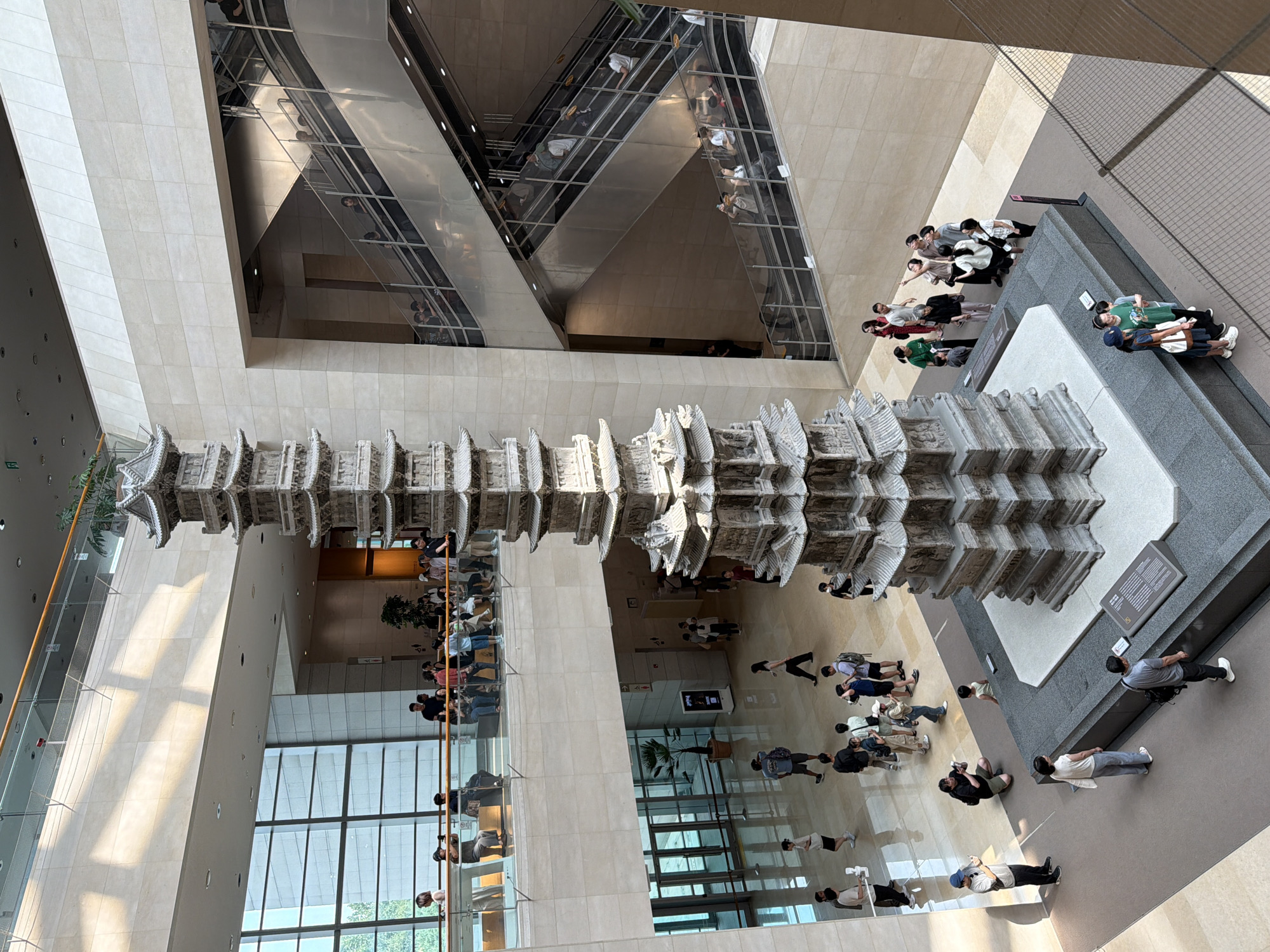 The image shows the interior of a modern museum atrium in Seoul, South Korea, featuring a tall, multi-tiered stone pagoda as the central exhibit. The pagoda is intricately carved and sits on a dark pedestal. Several levels of balconies and escalators are visible, with natural light streaming in from large windows.

Numerous visitors are scattered throughout the space, observing the pagoda or moving between levels. On the ground floor, people are seen standing, walking, and taking photographs. One person is kneeling to photograph the pagoda. On the upper levels, groups of people are gathered on balconies and riding escalators.

The architecture of the museum is characterized by light-colored stone walls, glass balustrades, and modern escalators. The overall atmosphere appears to be one of quiet observation and cultural engagement.