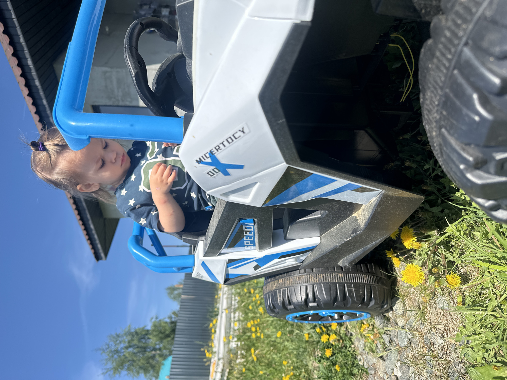 A young child with blonde hair sits in a white and blue toy buggy, possibly a battery-powered ride-on vehicle. The child is wearing a blue shirt with white stars. The buggy is positioned on a grassy surface interspersed with yellow dandelions. A house with a gray roof is visible in the background, along with a fence made of corrugated metal. The location appears to be outdoors on a sunny day. There is no indication this image was taken in the city of Kharnavy, Russia.