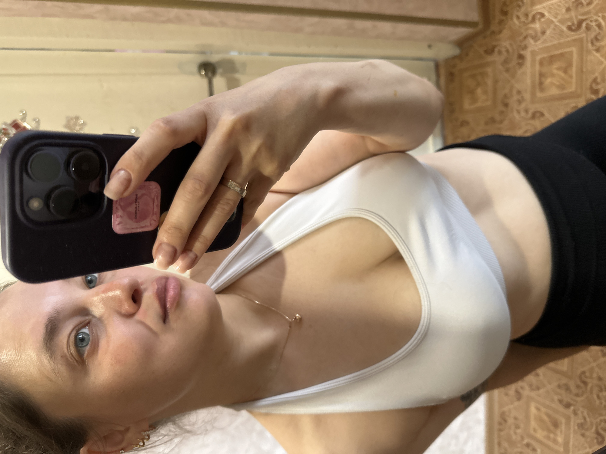 A young woman with light brown hair tied back is taking a mirror selfie in a domestic setting. She is wearing a white sports bra and black leggings. She has blue eyes and is wearing hoop earrings and a delicate necklace with a heart pendant. Her right hand, adorned with a ring, is holding a dark phone with a pink sticker on the back. The background shows a wall and a doorway, with a patterned floor visible. The location is identified as Yaroslavka, Russia.