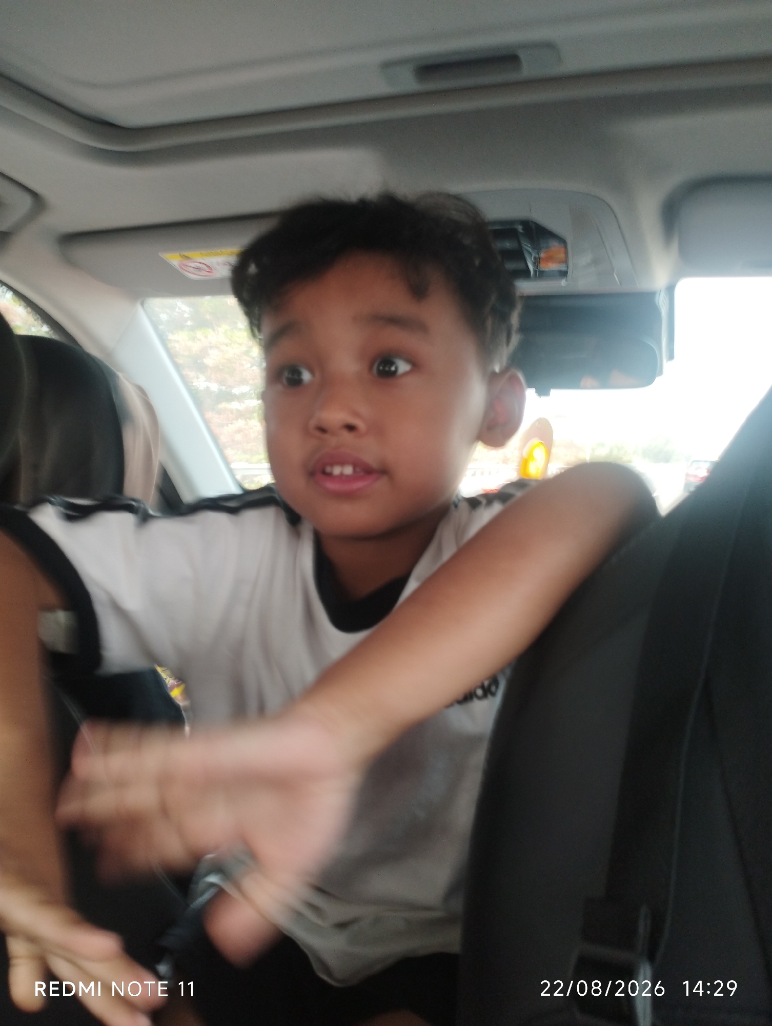 In the city of Cikampek, Indonesia, a young boy with dark hair is seated in a vehicle. He is looking to his right with wide eyes, his mouth slightly open. The boy is wearing a white t-shirt with black trim around the collar and sleeves. His right arm is resting on the back of a dark-colored seat, and his left hand is blurred due to motion, suggesting he is actively moving. The interior of the vehicle is visible, including the sunroof, a rearview mirror, and the headliner. Outside the vehicle, through the windows, trees and other vegetation can be seen. A date and time stamp at the bottom right of the image reads "22/08/2026 14:29", and at the bottom left is "REDMI NOTE 11".