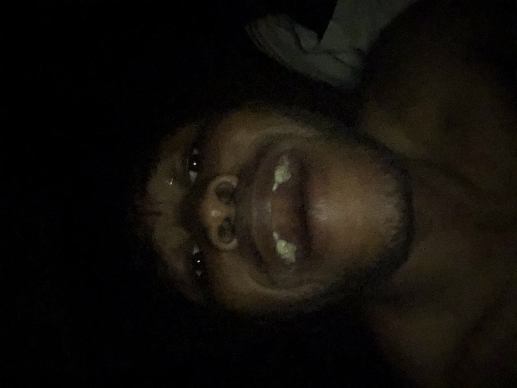 A close-up, poorly lit selfie shows a dark-skinned person with facial hair. The subject's mouth is open, revealing teeth. The setting appears to be indoors at night, although the location within Unknown, Unknown cannot be determined from the image.