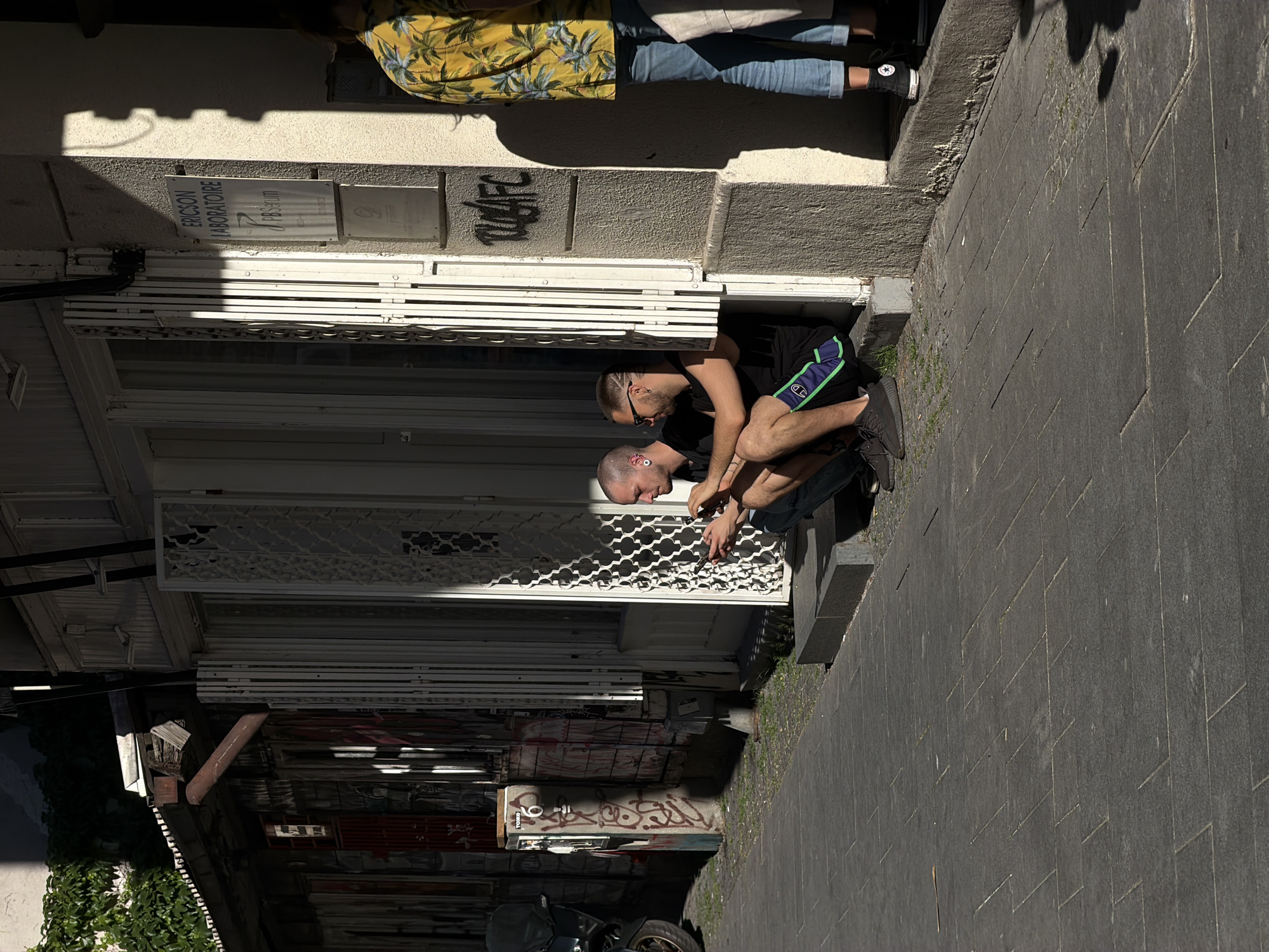 Two men are kneeling in a doorway, appearing to work on or repair a metal security gate. The younger man is wearing black shorts with green trim and a black t-shirt. The older man is wearing jeans and a dark-colored shirt. They are using tools to manipulate the gate. The doorway is set into a building with graffiti on the wall. A street with dark gray paving stones extends from the doorway. A person wearing a yellow floral shirt and jeans is standing on the sidewalk in the background. A sign on the wall reads "ERICSON LABORATOIRE PBS serum". Based on the sign and architectural style, this is in Belgrade, Serbia.