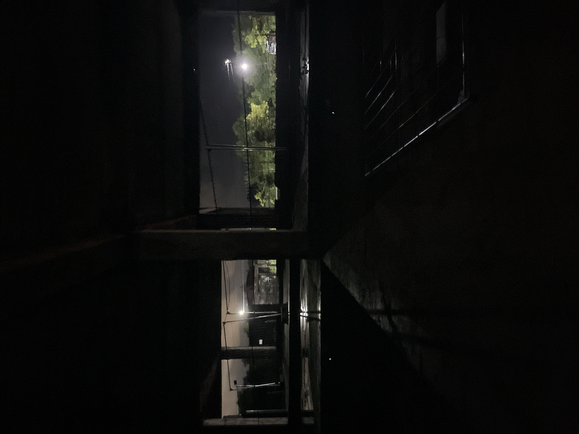 The image depicts an interior view of an unfinished concrete structure at night, likely in Poti, Georgia. The foreground is dominated by darkness, with a wet concrete floor reflecting faint light. In the mid-ground, large concrete pillars support the structure, creating dark, shadowed areas. Beyond these pillars, large openings reveal an exterior scene.
The exterior view shows a dimly lit landscape with trees illuminated by artificial lights. A tall lamppost with a bright light source is visible, casting a glow on the surrounding foliage. In the distance, the faint outlines of buildings and possibly vehicles can be discerned. The sky is dark and overcast, suggesting a nighttime setting. The overall impression is one of an abandoned or under-construction site, characterized by its stark architecture and nocturnal illumination.