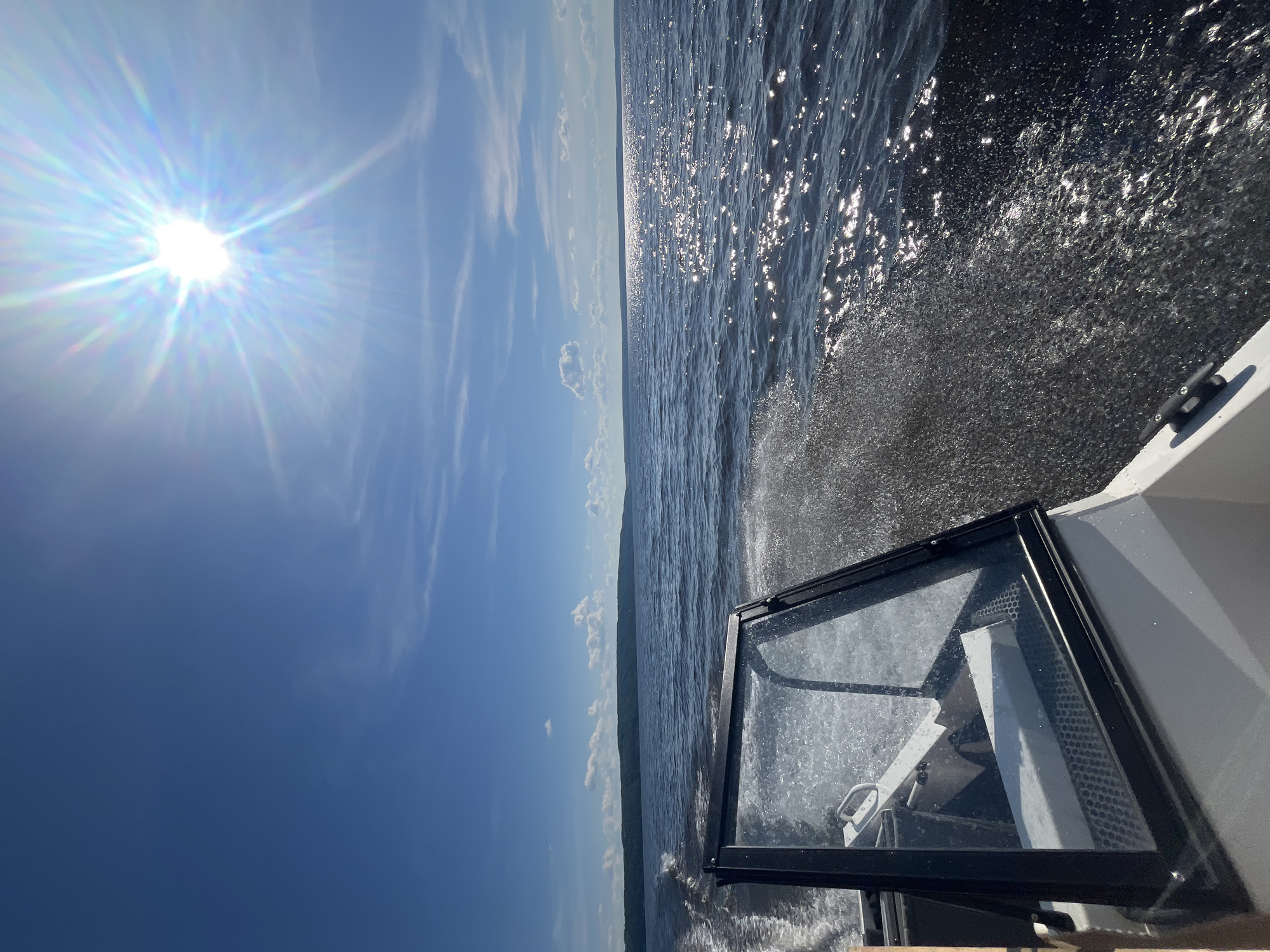 The image depicts a perspective from a boat moving on a body of water. The scene is dominated by the bright sun, which is high in the sky, suggesting it is daytime. The sun's rays are spread out, creating a lens flare effect across the blue sky. Wispy clouds are scattered across the sky. The water is choppy, with white spray coming off the bow of the boat as it cuts through the waves. The sun's reflection glints off the surface of the water, creating a sparkling effect. The horizon line is visible in the distance, showing a faint outline of land. The foreground features the edge of the boat, including a windshield with a black frame, and part of the interior, which appears to be a helm. The overall mood is one of speed and exhilaration on a sunny day. The location is identified as Siukterka, Russia.