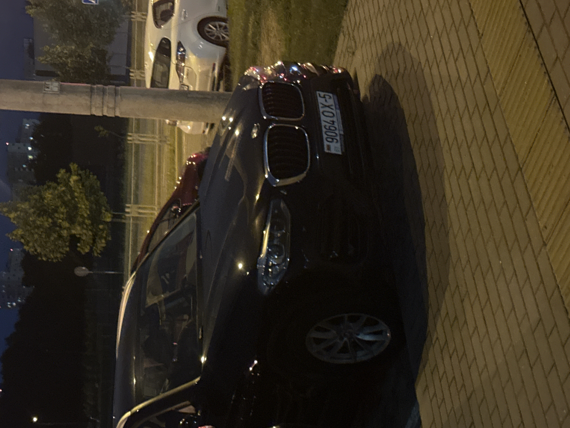 The image shows a black BMW parked on a paved area at night. The car's license plate reads "9064 OX-5". In the background, there are trees, streetlights, and buildings, indicating an urban or suburban environment. A white car and a red car are also partially visible. The paved area appears to be a sidewalk or parking lot, with a distinct tactile paving strip at the bottom right. The overall lighting suggests it is nighttime. The location is identified as Baravliany Village Council, Belarus, likely a street or public space within this area.