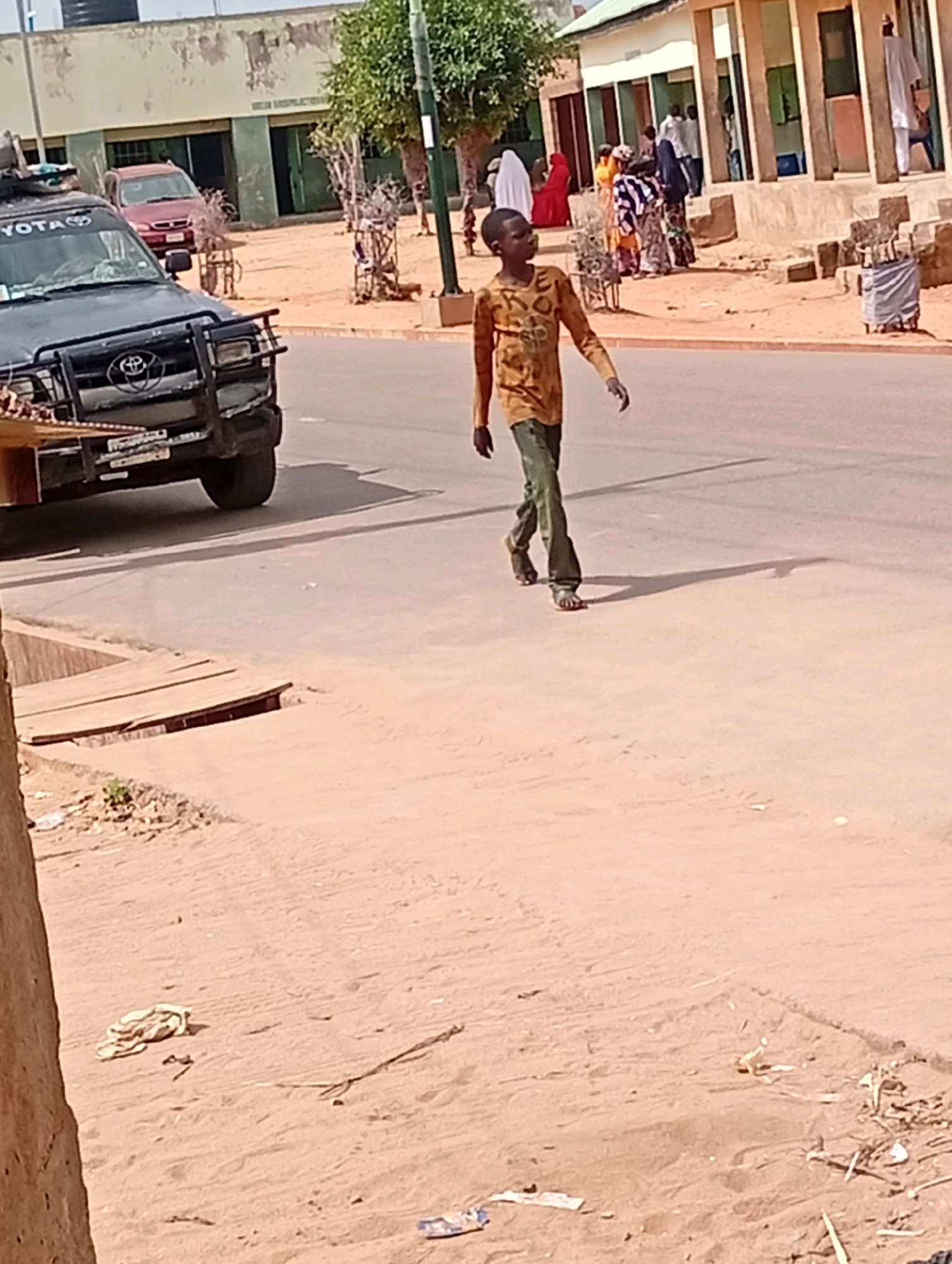 A young boy in a yellow, long-sleeved shirt and green pants walks across a dusty, paved road in Gombe, Nigeria. He is walking from right to left in the frame, facing away from the camera. In the background, several buildings with light-colored walls and dark trim are visible. People in various traditional clothing are gathered outside one of the buildings. To the left of the boy, a dark-colored SUV is parked on the side of the road. The environment appears dry and arid, with loose sand and debris on the sides of the road. A storm drain with a wooden cover is visible in the foreground on the left side of the image.