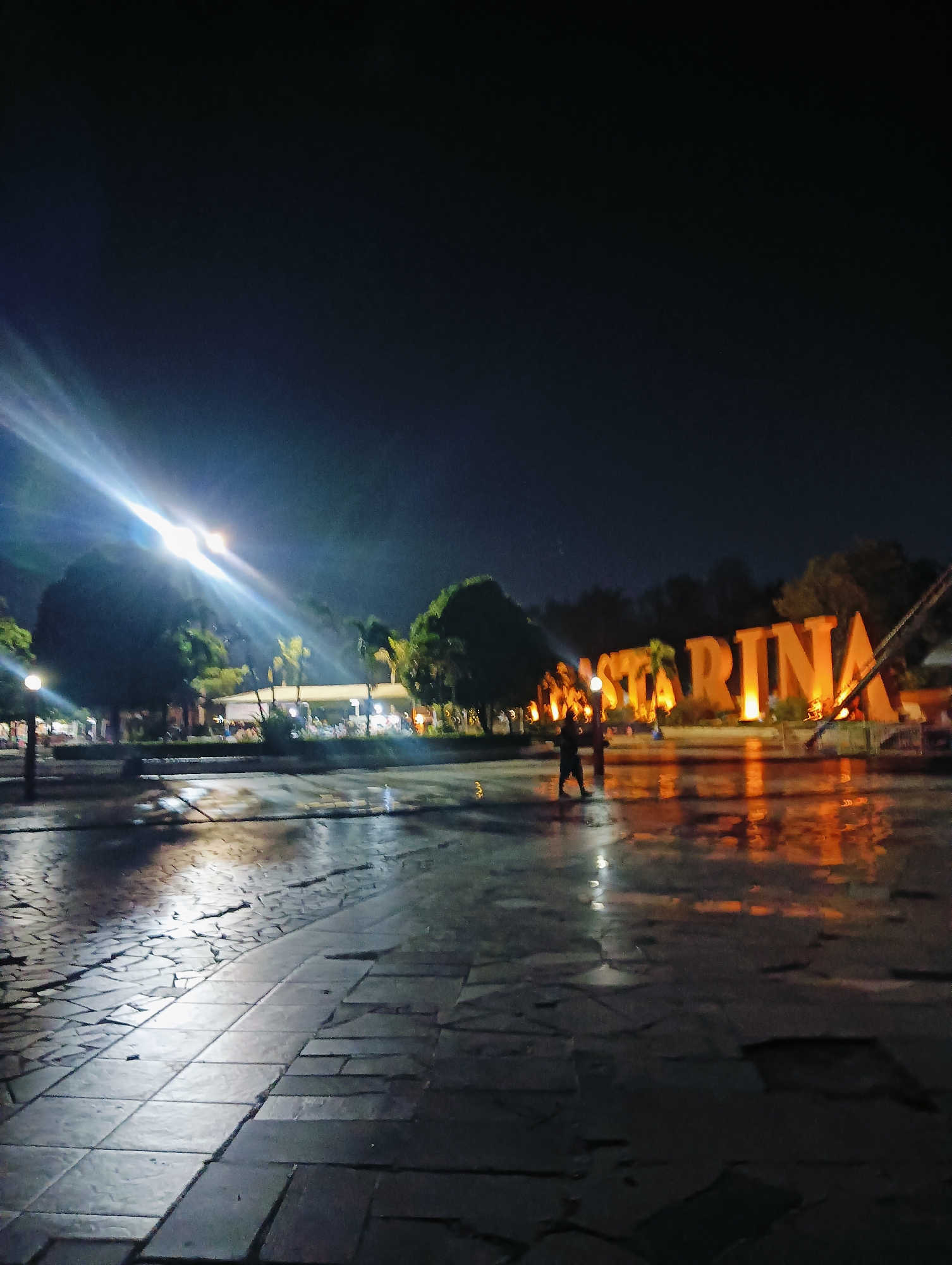 An exterior, eye-level, night-time photograph captures the "Bastarina" sign, illuminated with bright orange lighting, at the Batam City Waterfront, Batam, Indonesia. The sign is positioned in the background, with its large, block-style letters casting reflections on the wet, tiled ground in the foreground. A bright, white light beam from a light source on the left pierces the dark sky, creating lens flare and illuminating the left side of the scene. Two silhouetted figures are visible near the center of the frame, walking on the wet pavement. Trees and a building with additional lighting are present behind the sign, adding to the nighttime ambiance of the waterfront area.