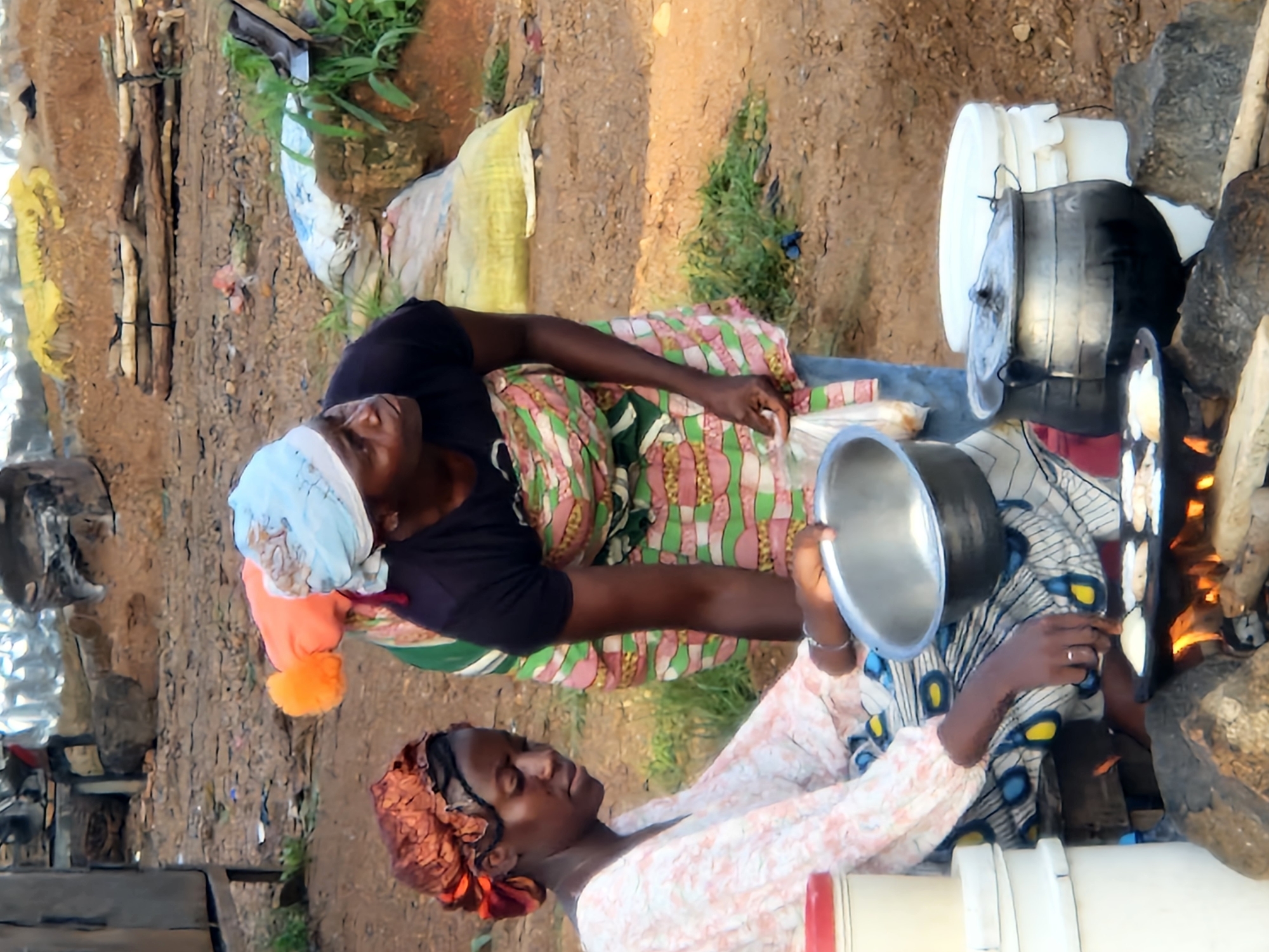 The image depicts an outdoor scene in Kabong, Nigeria, where two women are preparing food over an open fire. In the foreground, a woman wearing a patterned headscarf and a light-colored, floral-print top is seated. She is actively engaged in cooking, holding a metal bowl in her right hand and tending to small, flat, pale-colored items cooking on a griddle over flames. Beside her, to her left, are stacked white containers with red lids.
Behind her, another woman, also wearing a headscarf and a patterned apron over a dark top, stands and observes. Her hands are partially visible. The cooking area features a griddle placed on rocks over burning wood. To the right of the standing woman, large metal pots are visible, one with a lid.
The environment appears to be a dusty, unpaved area with sparse vegetation. In the background, there are piles of dirt, what appear to be logs or building materials, and large sacks, suggesting a possibly industrial or market setting. The overall impression is one of daily life and food preparation in an outdoor, possibly informal, setting.