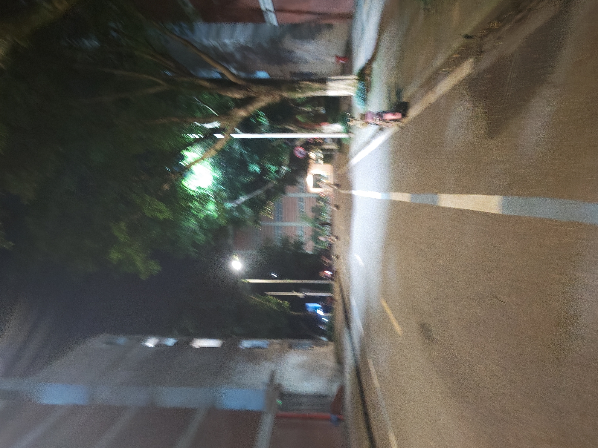 The image depicts a blurred nighttime street scene. A multi-story building, possibly an apartment complex or dormitory, is visible on the left side of the street, featuring multiple windows. Across the street, another building is partially visible on the right, with trees lining the sidewalk. A street lamp illuminates the area. A pink-colored scooter is parked on the right sidewalk. Several figures, appearing as blurred shapes, are present in the distance, some near the scooter and others further down the street, suggesting pedestrian or vehicular activity. The road is paved with asphalt and marked with white and yellow lines. The overall impression is of a street in a residential or campus area at night, with the blur indicating motion or low light conditions.