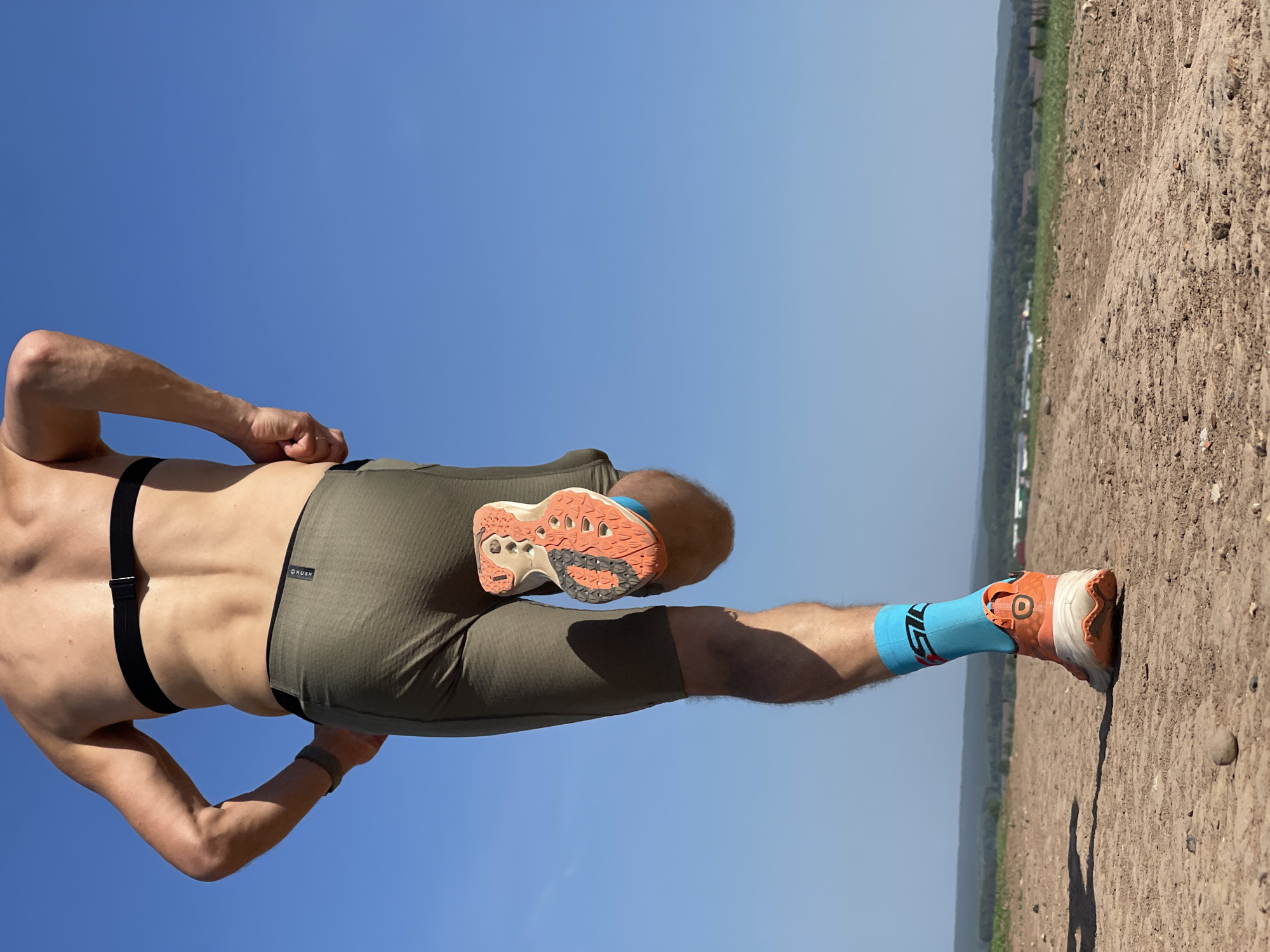 A man is captured mid-stride running along a dirt path in Perevoznoe, Russia. He is wearing olive-green running shorts, a black heart-rate monitor strap, and a sports watch. His running shoes are orange and white, paired with bright blue socks. The foreground shows the dirt path, while the background features a treeline and a hazy sky. The sky above is clear and blue.