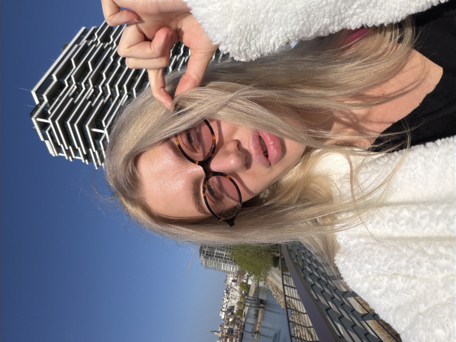 A blonde female subject is depicted in a close-up selfie, wearing oval, tortoise-shell patterned glasses with reddish-tinted lenses and a white textured sherpa jacket over a dark garment. Her left hand, with fingers extended, pinches a strand of her blonde hair near her temple. The background reveals a clear blue sky. To the subject's right, a distinctive modern high-rise building, Kula Belgrade (Belgrade Tower), featuring a white, layered, and stepped facade with dark windows, is prominent. To the subject's left, a metal railing and a concrete promenade overlook the Sava River. Across the river, a portion of the Belgrade cityscape, including various buildings and a distant spire, is visible. The scene is located along the Belgrade Waterfront in Belgrade, Serbia.