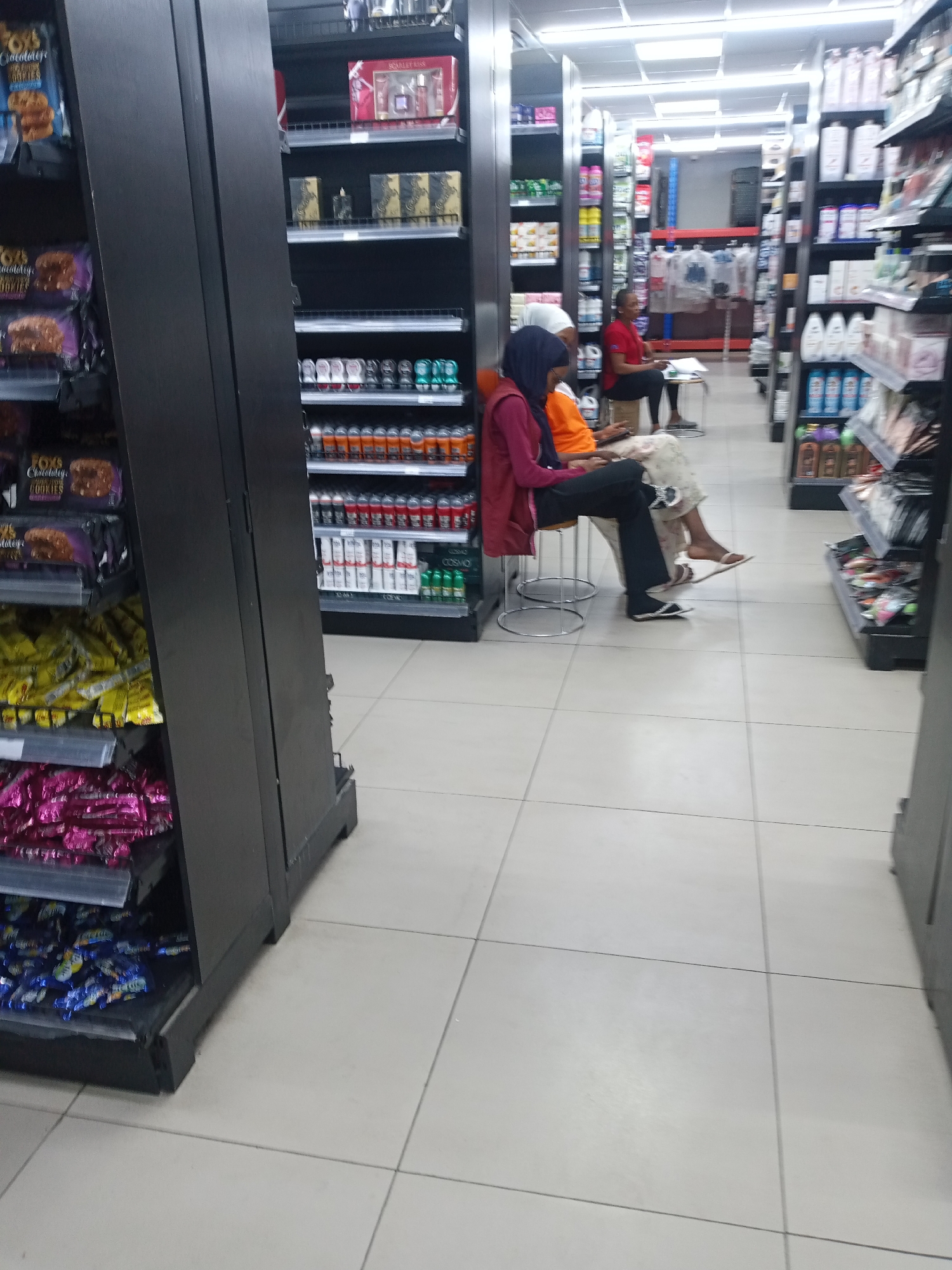 The image displays an indoor commercial retail establishment, located in Kawaji, Nigeria. The facility features aisles of uniformly black shelving units stocked with various consumer products, set on a floor of light-colored square tiles with dark grout lines. Overhead fluorescent lighting illuminates the entire space.
In the mid-ground, three individuals are seated on stools. On the left, a person, likely female, wears a dark blue hijab, a maroon long-sleeved top, and black trousers, seated on a metal stool and looking downwards. Next to her, a second person, also likely female, wears an orange long-sleeved top and a patterned lower garment, seated on a similar metal stool and looking downwards; she wears a white slip-on sandal on one foot, with the other foot appearing bare or in very light footwear. Further back and to the right, a third person, likely female, with short hair, wears a red short-sleeved top and dark trousers, seated on a low stool while appearing to engage with an item on a small white table positioned in front of her.
The shelving unit in the left foreground prominently displays multiple packages of "FOX'S Chocolately BIG CHUNKY COOKIES," along with shelves containing numerous packages of yellow, pink, and blue-wrapped items, consistent with snacks or confectionery. The shelving units in the right mid-ground are stocked with personal care products, including perfume gift sets, one labeled "SCARLET KISS," various boxed fragrances, and multiple rows of aerosol cans such as deodorants, with some products from the brand "COSMO" visible. The aisles extend into the background, revealing additional stocked shelving and a structural racking system in blue and red.