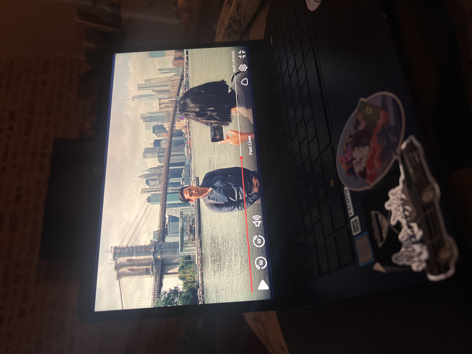 A laptop screen displays a show titled "Past Lives" featuring a man in front of the Brooklyn Bridge and the Manhattan skyline. A woman is also visible, holding a phone as if taking a photo. On the laptop's surface, several stickers are present near the keyboard. The laptop is positioned in a dimly lit room. The show is paused.