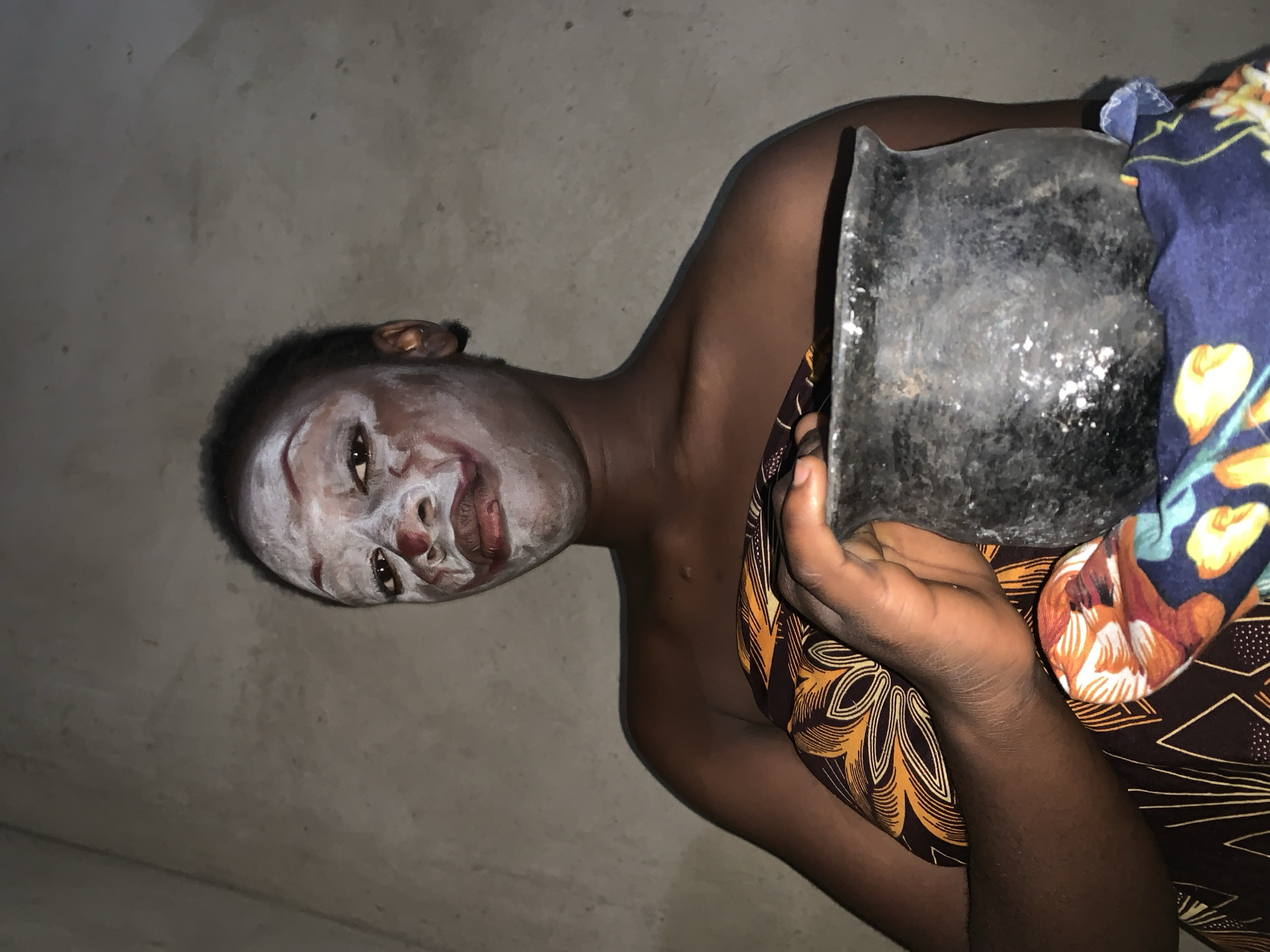 A person with white face paint, possibly indicating a ritualistic or ceremonial preparation, is holding a dark-colored, possibly metallic, vessel or object. The person has dark skin and is draped in patterned fabrics in yellow/brown and blue/orange/white. The person is located indoors, judging by the simple, uniform gray wall behind them. The location, Agban, Nigeria, is specified only as context, and no specific landmarks or identifying features are visible in the frame to confirm the location. The image is rotated, possibly to prevent automated bots from scraping the images or text from the image.