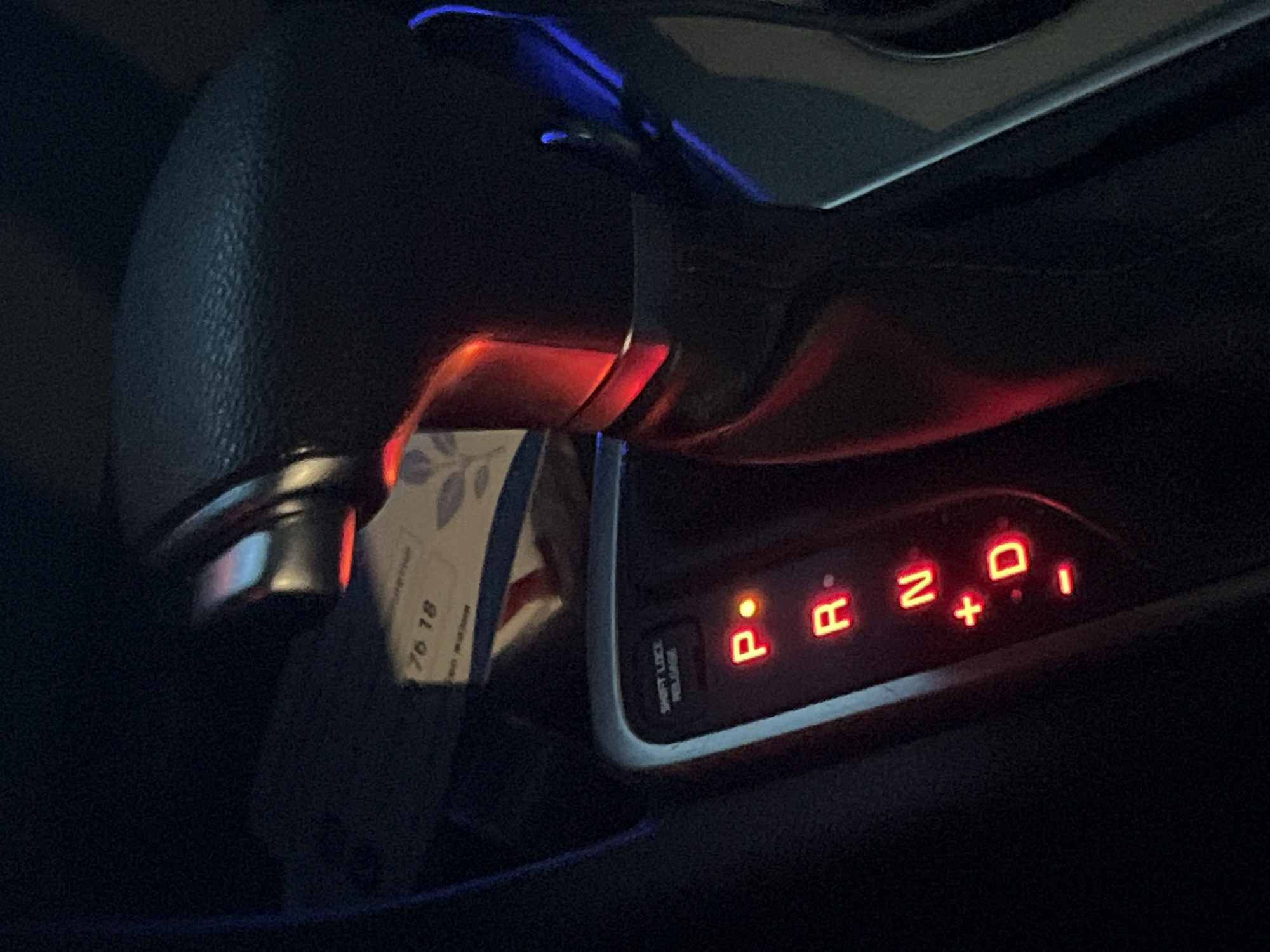 The image shows the gear shift of a vehicle in park (P). The illuminated letters "P R N D + -" are visible on the console. A piece of paper or card is tucked near the gear shift handle, with the numbers "76 18" visible. The vehicle's interior is dark, suggesting nighttime. The location is unknown and cannot be determined from the image. There is no evidence to suggest that this image was taken in Yerevan, Armenia.