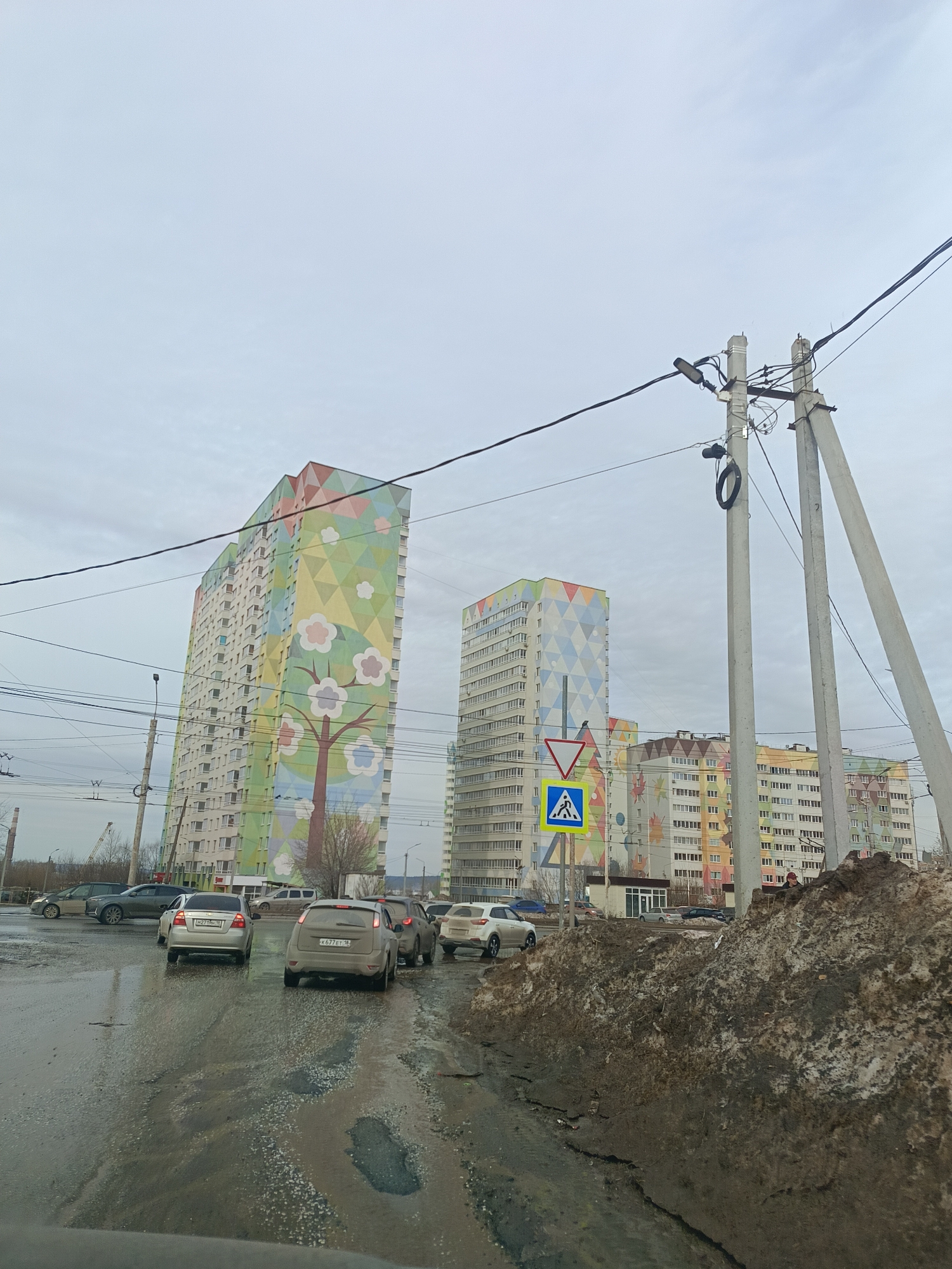 The image depicts a street scene in Izhevsk, Russia, identifiable by the distinctive, brightly colored facades of the apartment buildings in the background. The buildings feature geometric patterns and floral motifs. A row of cars is visible on a wet, potholed road, suggesting recent rain or melting snow. A pedestrian crossing sign is present next to a large pile of dirt and snow, indicating an urban environment. Concrete utility poles with power lines crisscross overhead. The sky is overcast, creating a dull, gray light.