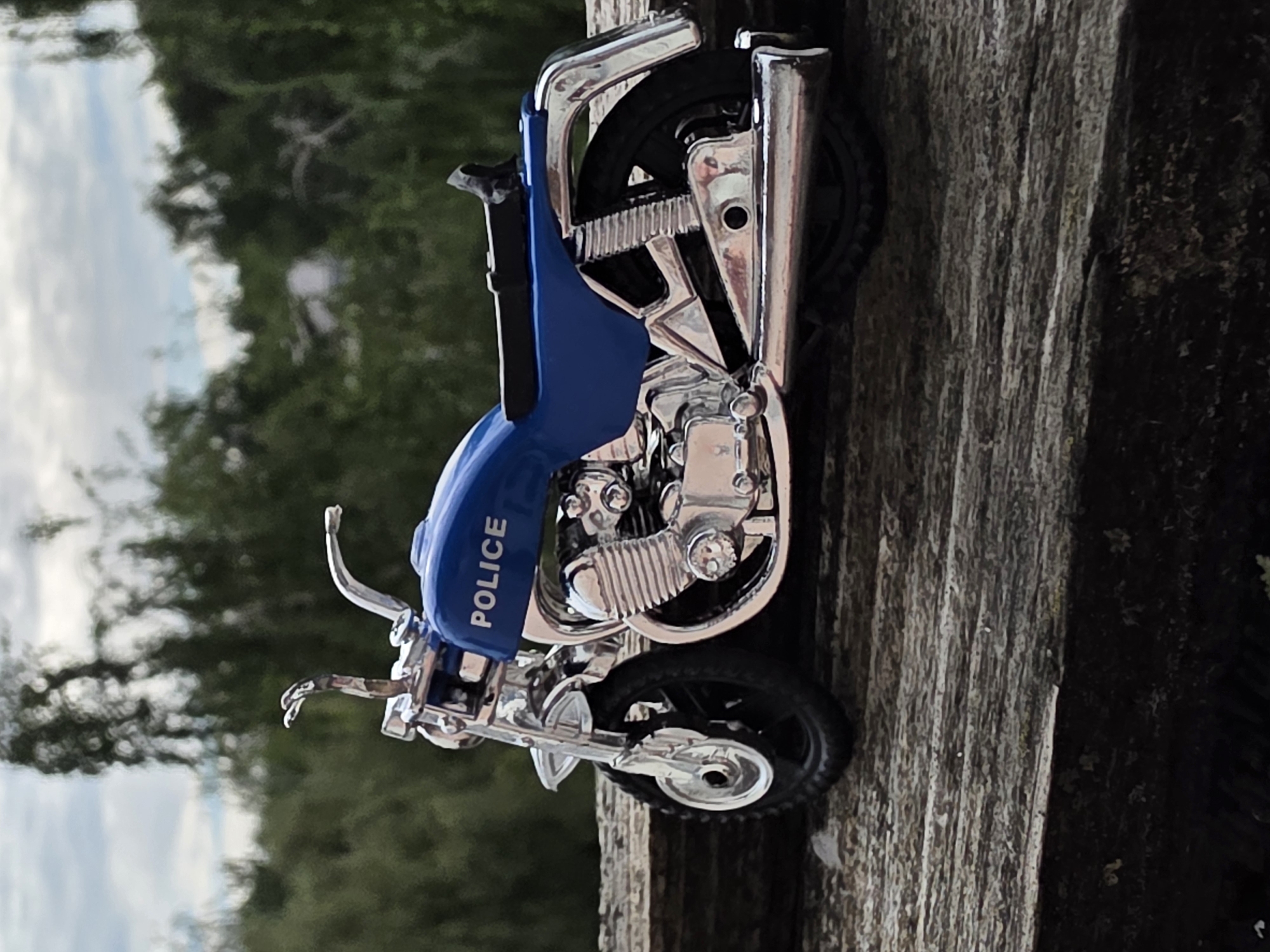 A blue and chrome toy police motorcycle is centrally positioned on a weathered wooden surface in Unknown, Unknown. The miniature vehicle faces left, with its right profile prominently displayed. The fuel tank and rear fender are a vibrant blue, featuring the word "POLICE" in white capital letters on the tank's side. Extensive chrome plating is visible on the handlebars, front forks, engine block (detailed with cooling fins), exhaust pipe, rear shock absorbers, and elements of the wheels and rear fender. A black seat is mounted above the rear wheel. The textured wooden surface beneath the motorcycle exhibits distinct grain patterns, horizontal lines, and areas of discoloration, suggesting age and outdoor exposure. The background is rendered in a soft-focus bokeh, showing blurred green foliage from trees or bushes beneath a light, overcast sky. The scene is illuminated by natural outdoor light, producing subtle reflections on the chrome components. No individuals or additional subjects are present.
