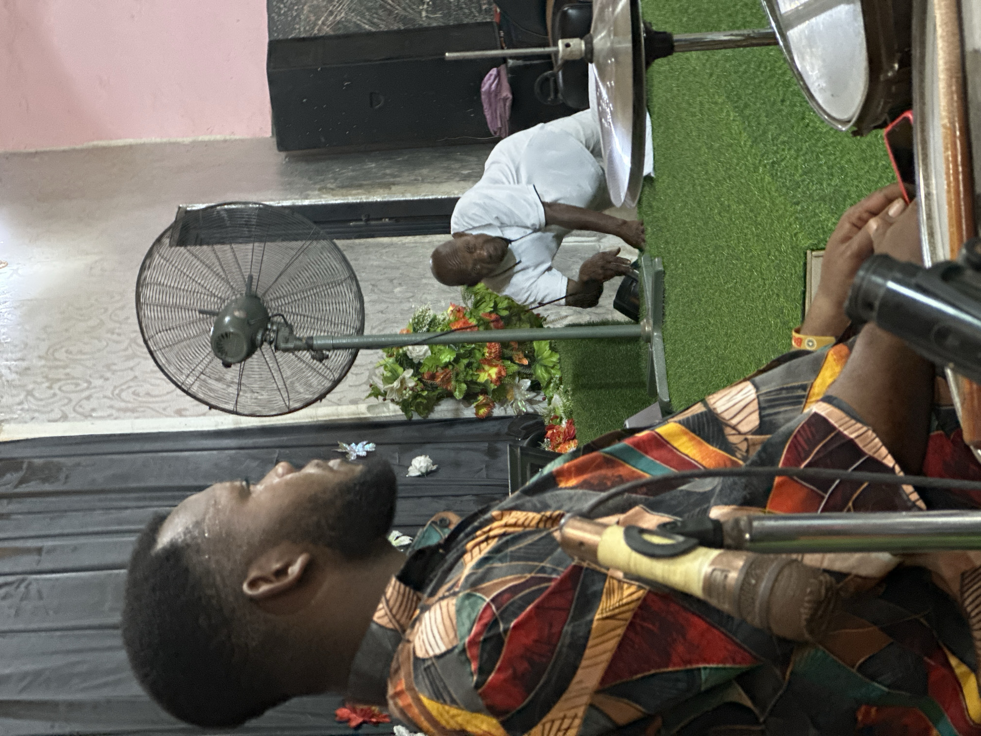 The scene takes place in what appears to be a room in Aba, Nigeria, as indicated by contextual clues. A man in a colorful patterned shirt and beard is positioned in the foreground, with his head tilted upwards. A microphone is visible near him, suggesting a possible performance or recording activity. Behind him, another man wearing a white shirt is kneeling, possibly interacting with some equipment or items on the floor, which is partly covered with a patch of artificial grass. A black fan and a flower arrangement are also present in the background. The room has a tiled floor and the walls are painted in light colors.