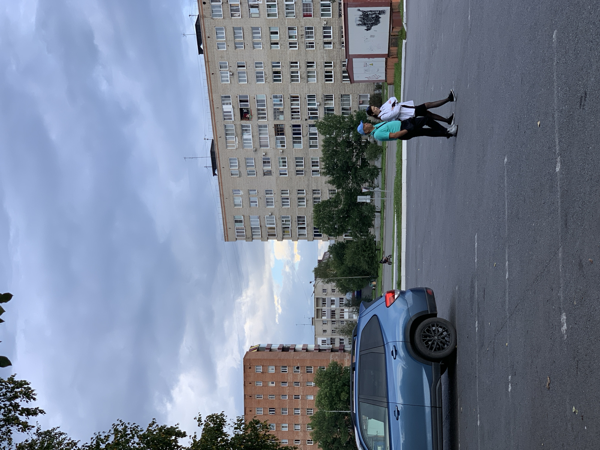 The image depicts an outdoor scene in the city of Prokopyevsk, Russia, featuring a parking lot, residential buildings, and two individuals. A blue SUV is parked in the foreground on the left. In the mid-ground, two people, a man wearing a turquoise shirt and blue cap, and a woman in a white jacket and dark skirt, are walking together from left to right. Behind them, several multi-story residential buildings are visible. The sky is overcast with clouds. A distant figure on a motorcycle can be seen further back. The scene appears to be in a residential area of the city.
