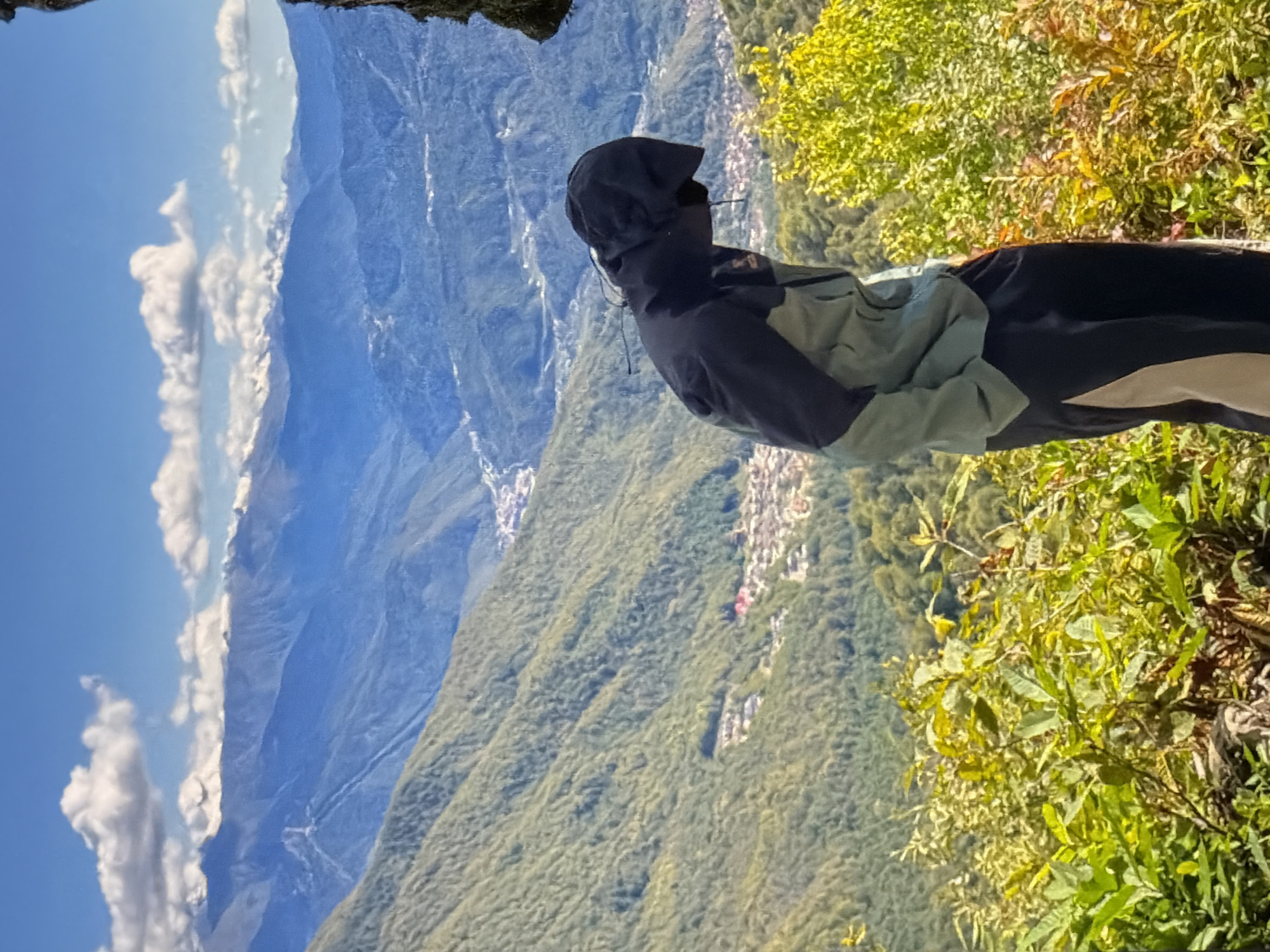 A person wearing a dark jacket with a hood stands on the edge of a cliff overlooking a mountainous landscape. The person is looking towards the mountains. The landscape includes a town nestled in the valley between the mountains and a blue sky with clouds. The exact location is Unknown, Unknown.