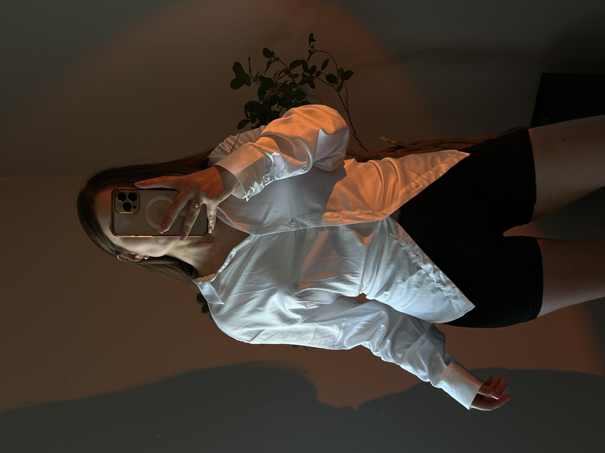The image is a selfie taken by a person in Ternopil, Ukraine. The person is wearing a white button-down shirt and black shorts. They are holding a smartphone in their right hand, using it to take the picture in a mirror. A plant with green leaves is visible behind the person. The lighting in the room is dim, with warm-toned light casting shadows. The overall mood appears to be relaxed and casual. The time of day is not explicitly indicated, but the lighting suggests it could be evening or indoors with artificial lighting. The background is mostly dark, with some hints of furniture or other objects not clearly discernible. There is no visible text in the image.