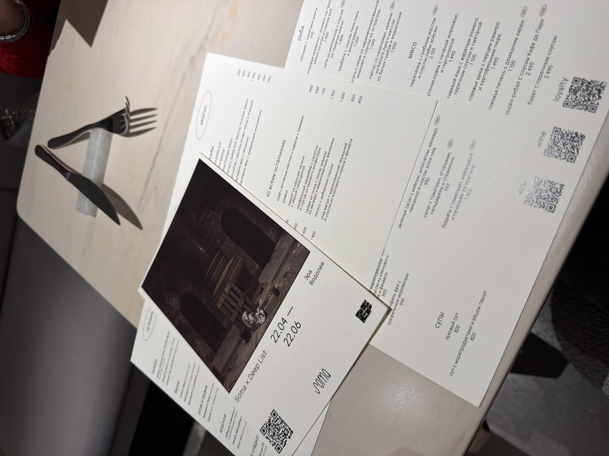Here is a detailed description of the image:
**Scene:** The image shows a restaurant table setting. A knife and fork rest on the table alongside a few sheets of paper. The papers appear to be a menu and a promotional flyer. The menu lists various food items such as appetizers, soups, fish, and meat dishes along with their prices. The flyer has the name "Soma x Deep List" and mentions dates (22.04 - 22.06) with an image and the name "Эра Водолея". The environment has a simple restaurant table surface.
**Moscow, Russia Location Inference:** The language on the menu and flyer is Russian. Considering this, it is likely that the restaurant is located in Moscow, Russia.