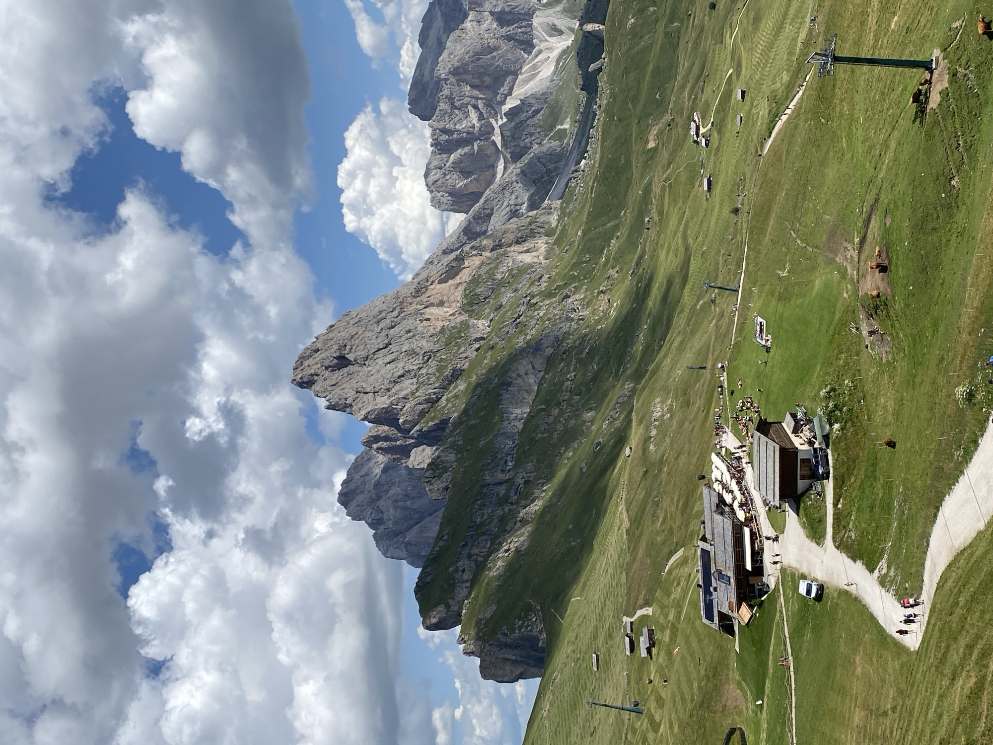 This image depicts a scenic mountain landscape in Santa Cristina Gherdëina, Italy, under a partly cloudy sky. In the foreground, a winding dirt path meanders through a vibrant green grassy slope, dotted with small wooden buildings. Several people are visible walking along the path, and a white van is parked near one of the buildings. Further up the slope, a larger complex of buildings with outdoor seating areas and umbrellas suggests a restaurant or mountain hut, with numerous people gathered. A ski lift tower stands prominently on the left side of the image, indicating a popular area for both summer and winter activities. In the mid-ground, the lush green mountainside is marked with terraced patterns. The background is dominated by a dramatic mountain range with jagged peaks and rocky formations, bathed in sunlight. A few cows are grazing on the grassy slopes. The overall scene captures a tranquil and picturesque mountain environment with evidence of human activity.