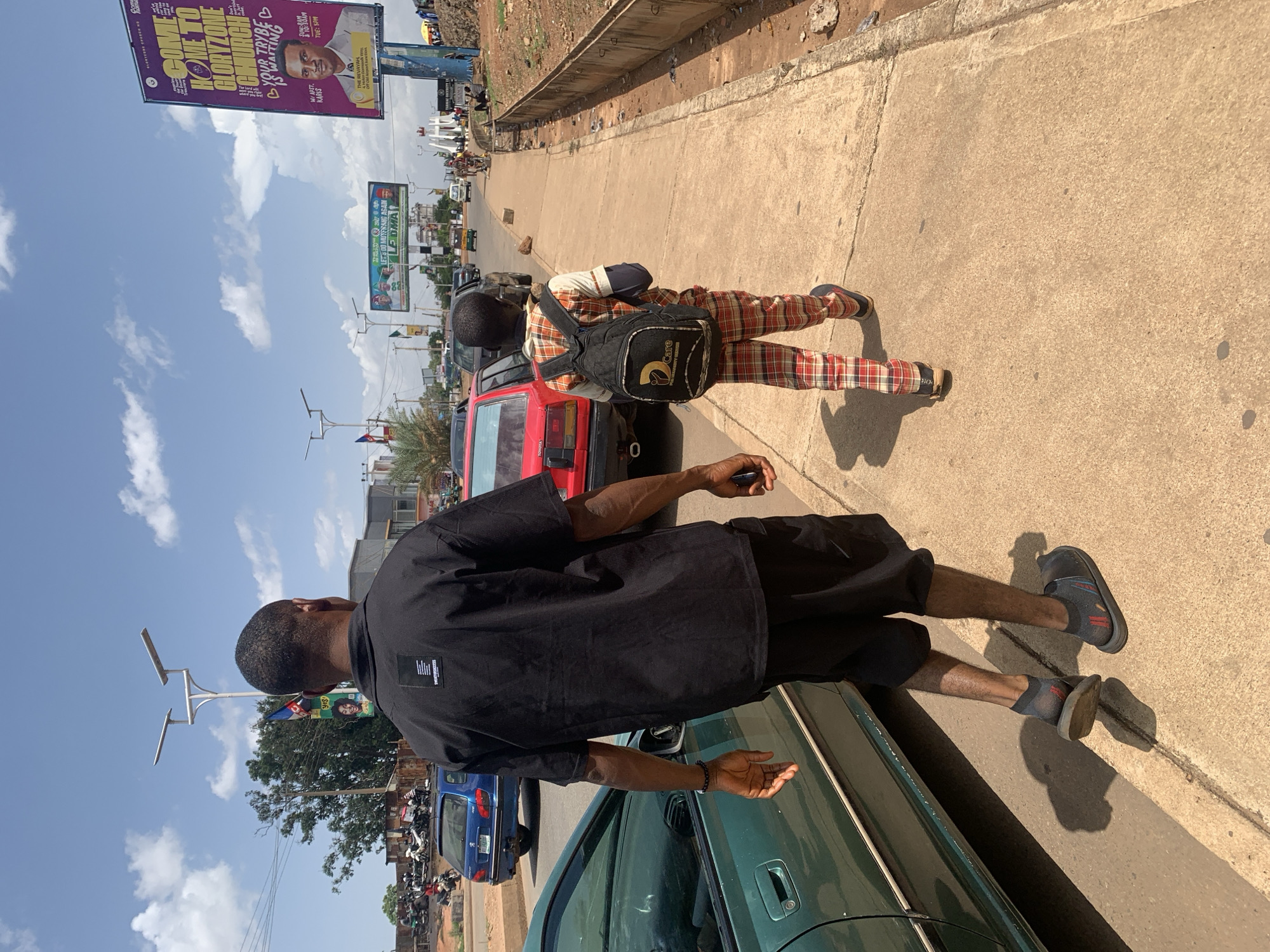 The image portrays a sunlit street scene in Jos, Nigeria, characterized by a tilted perspective. A clear blue sky with scattered white clouds dominates the upper portion.
In the foreground, a person, likely male, is partially visible from the back, wearing a black short-sleeved shirt and black shorts with dark sandals. He stands next to a green sedan, with his right hand possibly holding a phone. Further along the paved sidewalk, another person, also seen from behind, walks away from the camera. This individual wears a distinctive red, white, and black plaid shirt and trousers, carrying a black backpack adorned with a white "2Care" logo.
Parked cars, including a red Toyota sedan behind the plaid-clad person and a blue car further down the road, line the street. In the distance, more vehicles and motorbikes suggest an active urban environment. Modern streetlights with solar panels are visible along the road.
Two prominent billboards stand out in the background. The larger, purple one advertises "COME HOME TO GLORYZONE CHURCH" and states "YOUR TRYBE IS WAITING," featuring "W/ APST. KARIS" and "TUE: 5PM," alongside a portrait of a man and text including "THE REVAYAH." A smaller green billboard below it reads, "LETS DO MUYFWANG AGAIN" and "LEDMA." The strong shadows indicate bright daylight, likely midday or early afternoon. The scene captures typical daily activity on a bustling street in Jos.