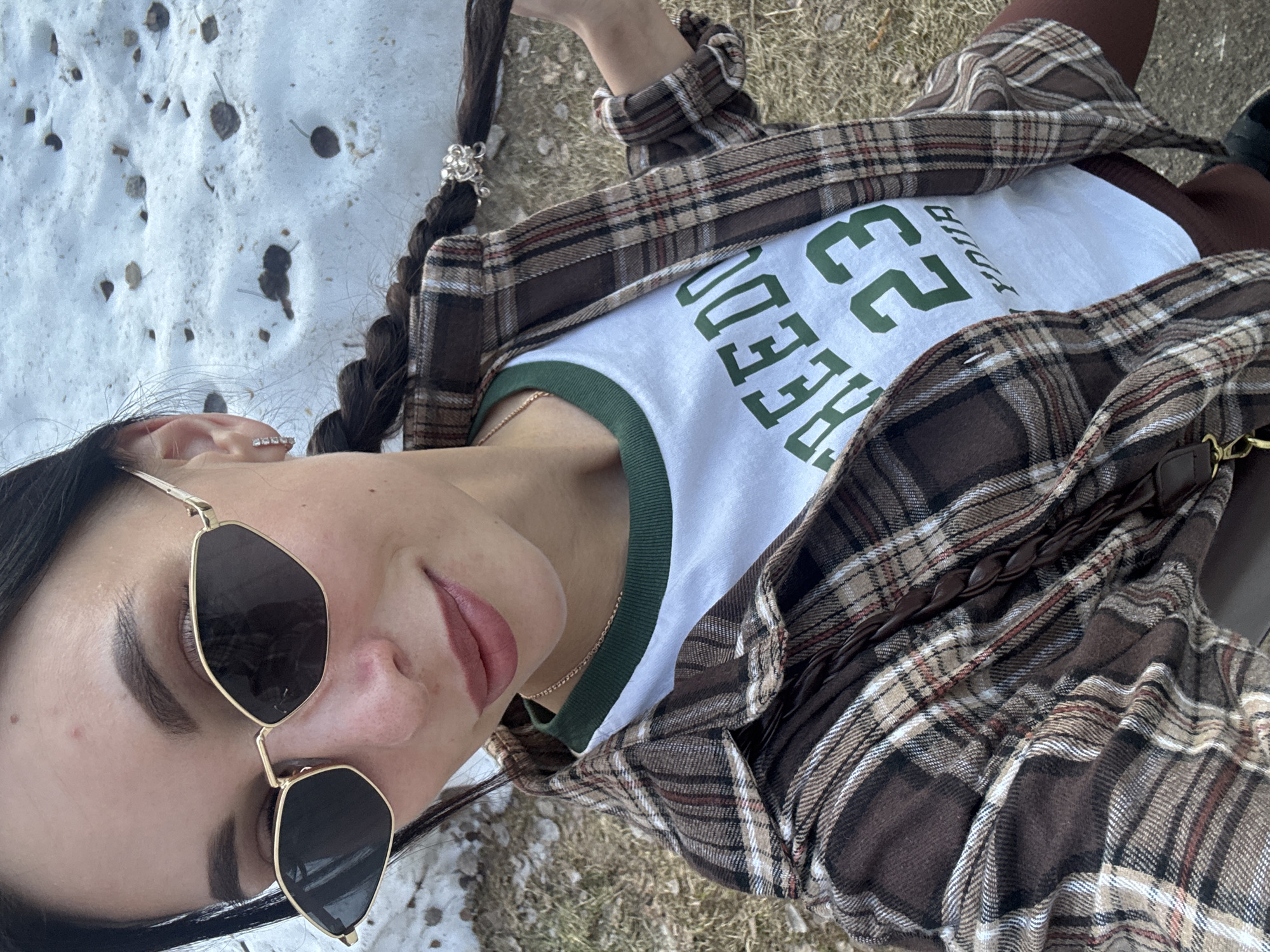 A young woman is pictured in a close-up, selfie-style shot. She has long, dark hair styled in a braid with a decorative silver clip, and she is wearing sunglasses with a geometric frame. She is dressed in a white t-shirt with green lettering, a plaid shirt, and brown pants. The background appears to be a combination of snow and grassy areas. The location, Noviy, Russia, is not discernible from the image.