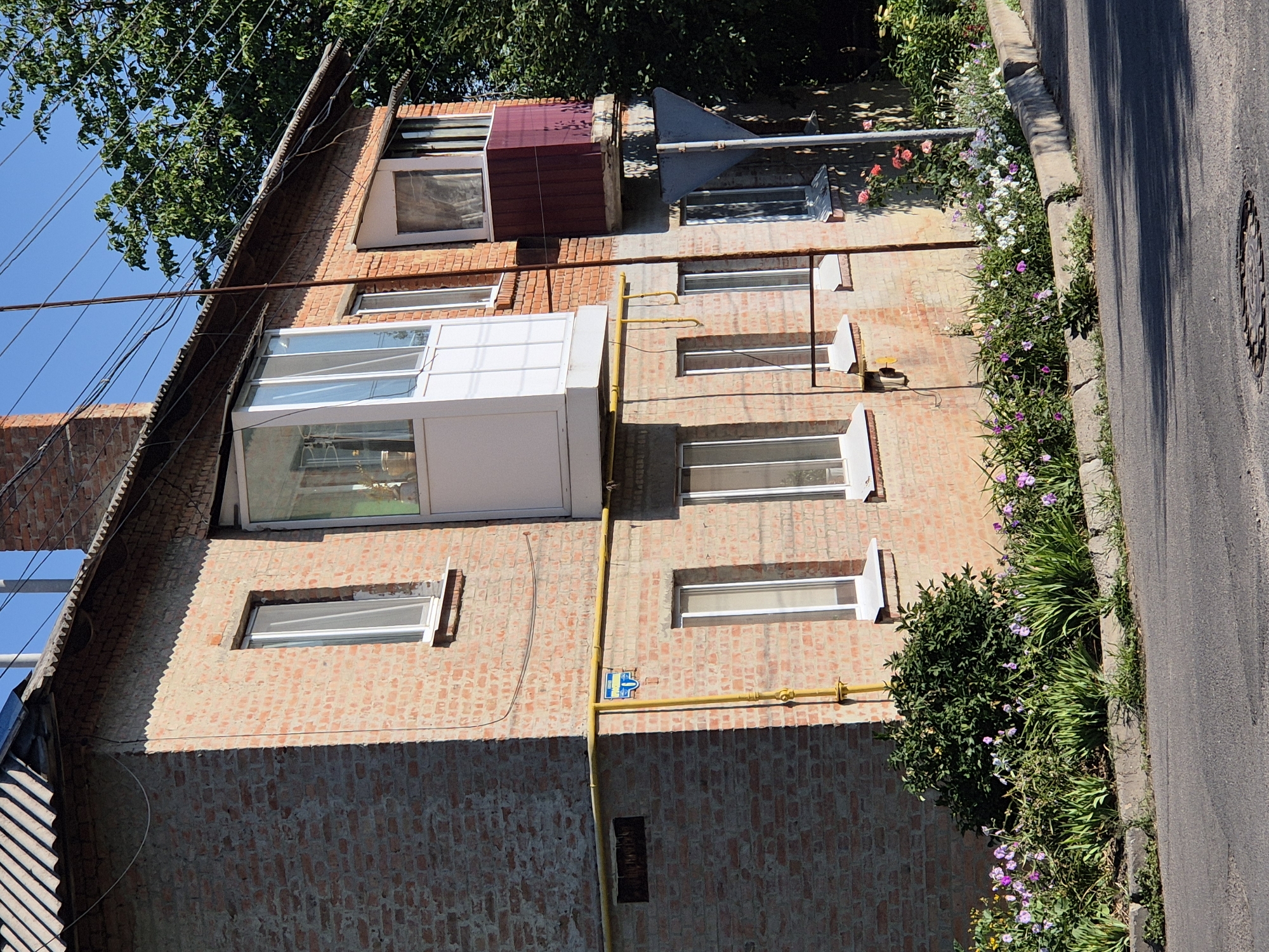 The image depicts a brick building located in Sozonivka, Ukraine. The building has multiple windows and two balconies, one of which is enclosed with glass. A yellow pipe runs along the exterior of the building, with a blue and white sign attached to it. In front of the building, there is a flowerbed with various types of flowers, including pink, purple, and white blooms, as well as green foliage. A concrete curb separates the flowerbed from the asphalt road. Trees are visible in the background. The sky is clear and blue. The scene is illuminated by sunlight, casting shadows on the ground.