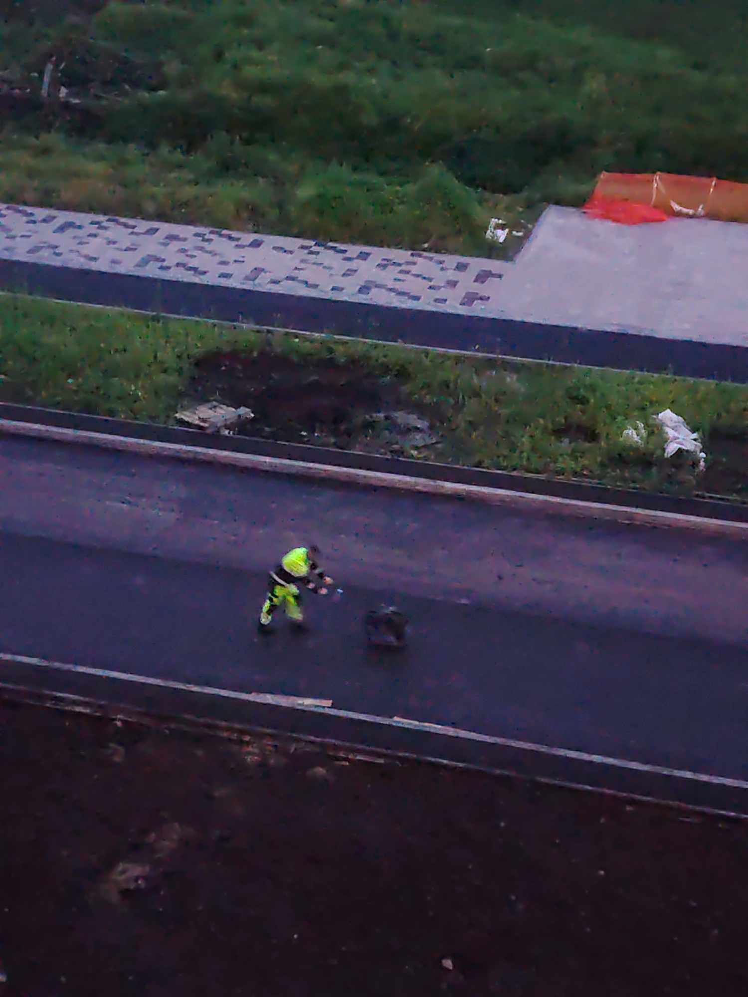 An aerial view shows a person in a high-visibility yellow and green outfit working on an asphalt road. The individual is holding what appears to be a tool or equipment in their hands. To the right of the worker, a dark, circular object is visible on the road. Adjacent to the road is a grassy area with some debris, and further beyond is a paved walkway with a pattern of darker squares. In the background, a large area of green vegetation is visible. The scene appears to be an outdoor construction or maintenance site in Yekaterinburg, Russia.