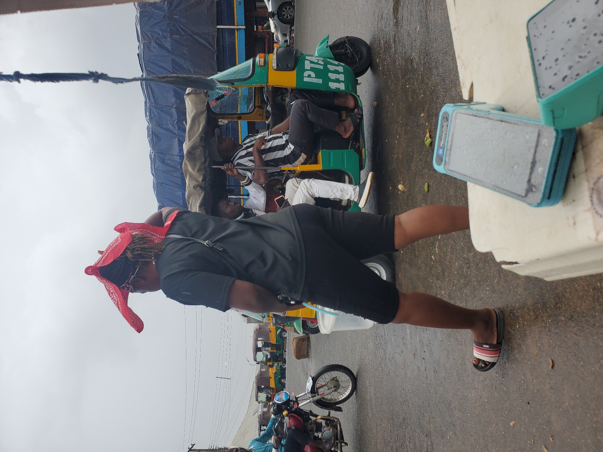 The image depicts a street scene in Gwong, Nigeria. The focal point is a person wearing a black t-shirt, black shorts, a red bandana on their head, and sandals. Their right leg is extended, and they are looking downwards. Behind this person, a yellow and green auto-rickshaw, also known as a "keke," is visible. Two other individuals are seated within the keke. One is wearing a black and white striped shirt and black pants, holding a metal rod. The other is wearing a white shirt and pants. The keke has "PTA 1111" written on its front.
Further back, a line of similar auto-rickshaws is parked. In the foreground, to the right of the main subject, are two turquoise electronic devices, possibly mobile phones or payment terminals, with screens displaying a grid-like pattern and water droplets. The ground is wet, suggesting recent rain or a damp environment. The sky is overcast, indicating a cloudy day. The scene appears to be outdoors, possibly a market or a transportation hub, given the presence of multiple vehicles. The overall atmosphere seems casual, with individuals engaged in daily activities.