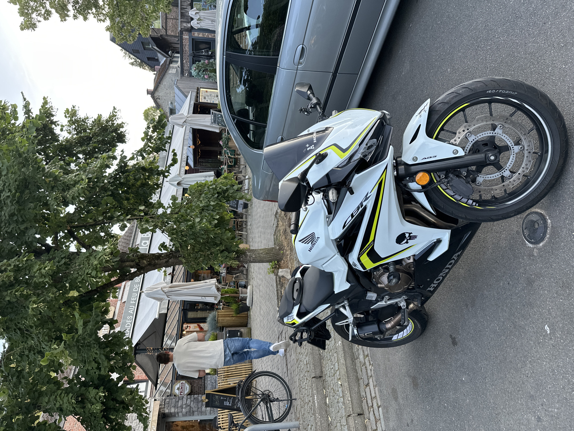 In Durbuy, Belgium, a white Honda CBR motorcycle with yellow accents and a skull decal is parked on a cobblestone street. The motorcycle is in the foreground, angled towards the right. A silver sedan is parked to the right of the motorcycle. In the background, a man wearing a light-colored t-shirt and blue jeans walks away from the camera. He is near a bicycle parked on the sidewalk. A building with an awning and outdoor seating is visible behind the man. Trees with green leaves are overhead, and the sky is bright, suggesting daytime.