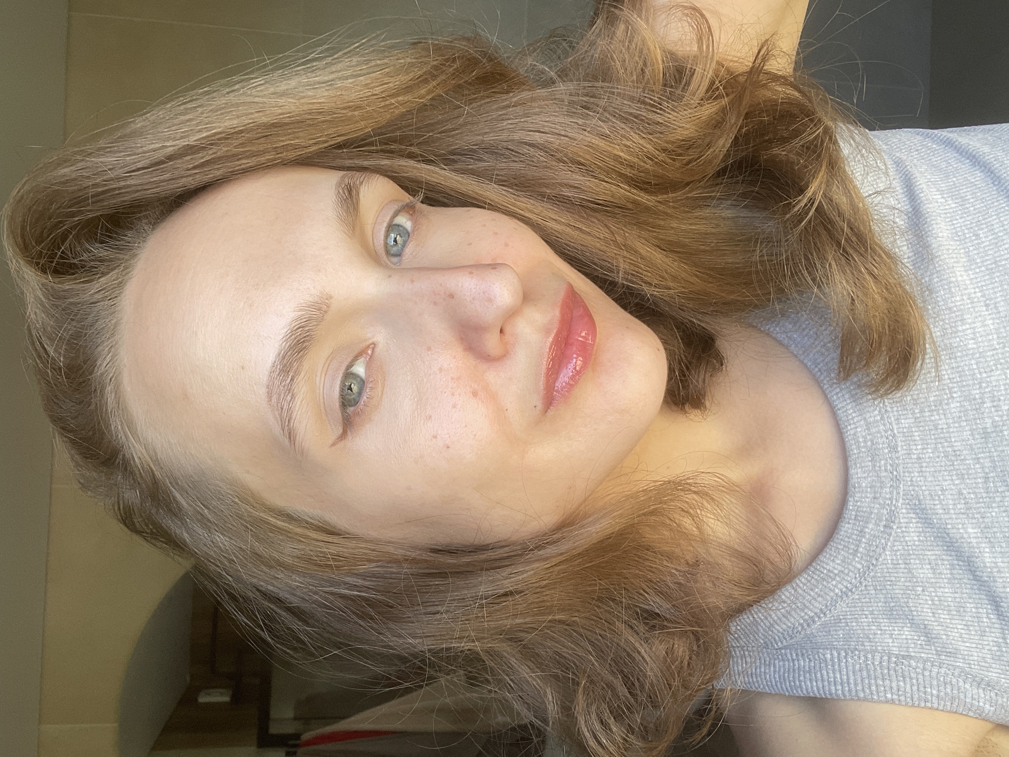 The image is a selfie of a woman with light brown hair, fair skin, and blue eyes. She has freckles on her face, particularly on her nose and cheeks. She is wearing a gray shirt. The background is blurry, but there is a hint of a doorway or archway to the left. There's no information to identify the location as Подшивалово, Russia.