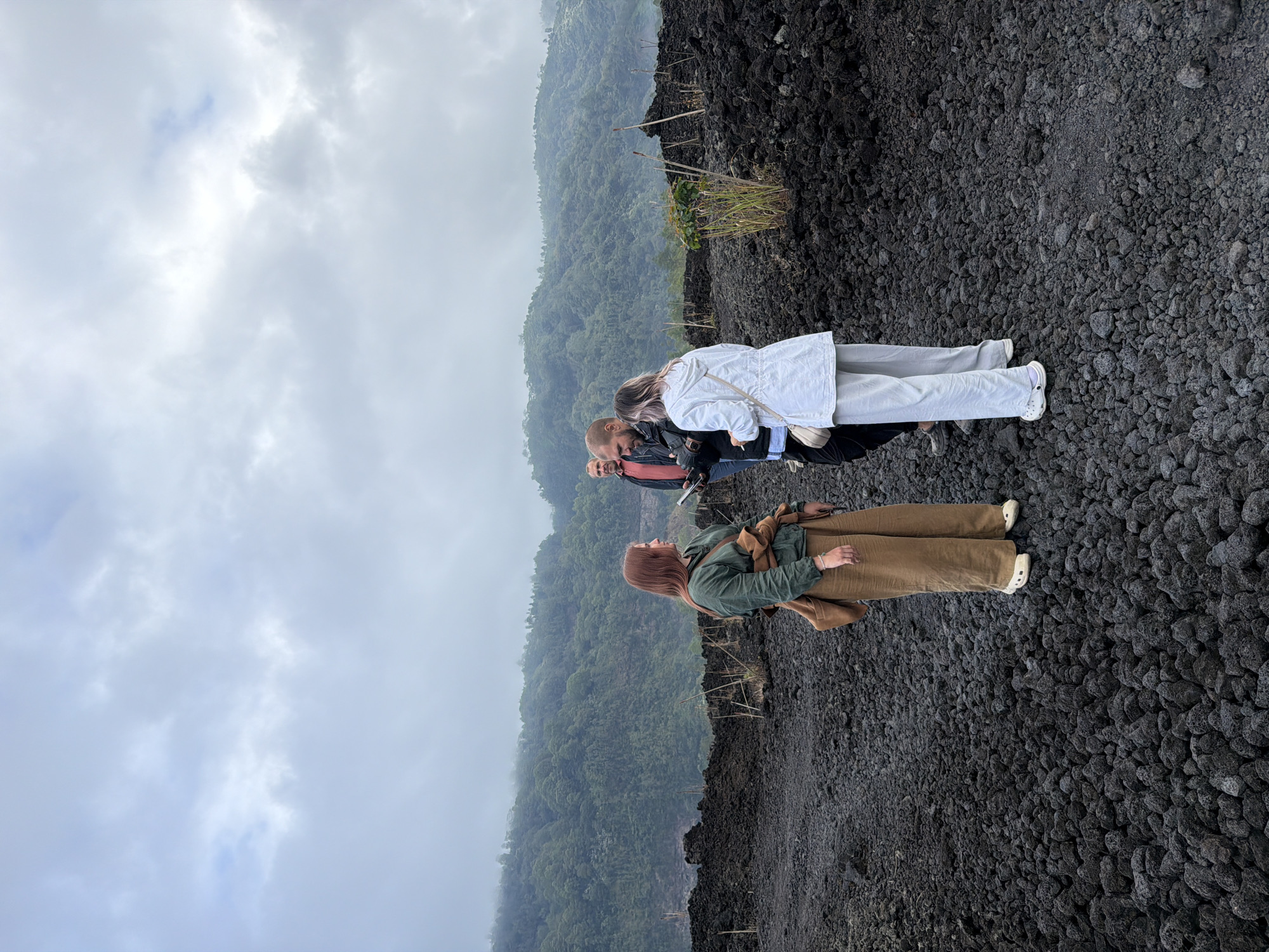 The image shows a group of four people standing on a landscape of dark volcanic rocks under a cloudy sky. In the foreground, a woman with red hair, wearing a green jacket and brown pants, is looking to her left. Beside her, a man in a black jacket and light-colored pants is looking at a phone. To their right, a woman in a white jacket and light-colored pants is facing away from the camera. In the background, a man in a red shirt stands behind the other three. The terrain is covered with small, dark, porous volcanic rocks. Beyond the rocks, a lush, green, forested mountainside rises up, shrouded in mist. The scene appears to be the rim of a volcanic crater or a recent lava field, consistent with the geological features found in Kintamani, Indonesia, a region known for its active volcanoes and crater lakes. The overall atmosphere is overcast and somewhat damp, suggesting a cool, possibly humid, climate.